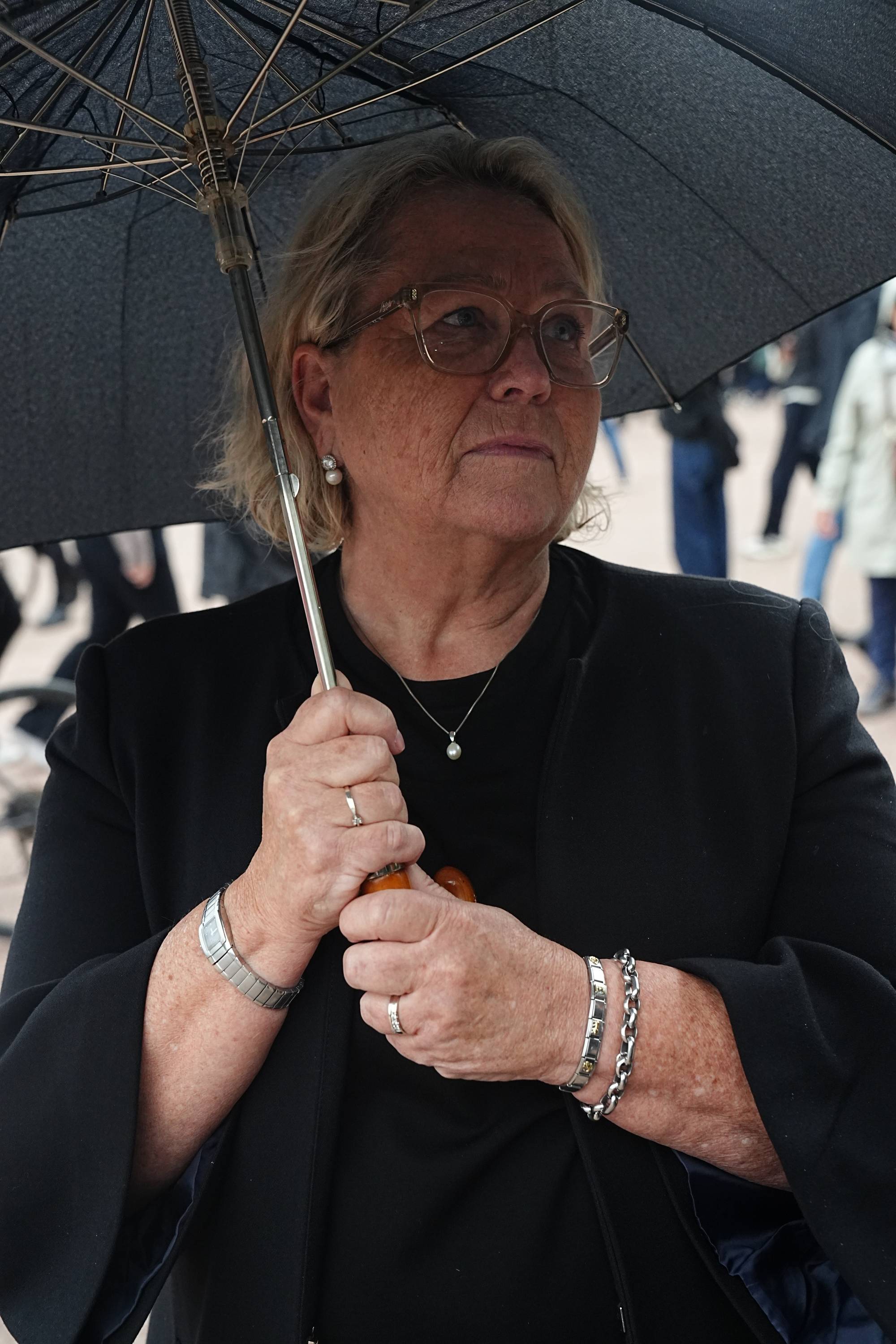 Kvinne med briller holder en svart paraply utendørs. Hun er kledd i en svart jakke og smykke. (Bildebeskrivelsen er laget av en KI-tjeneste)