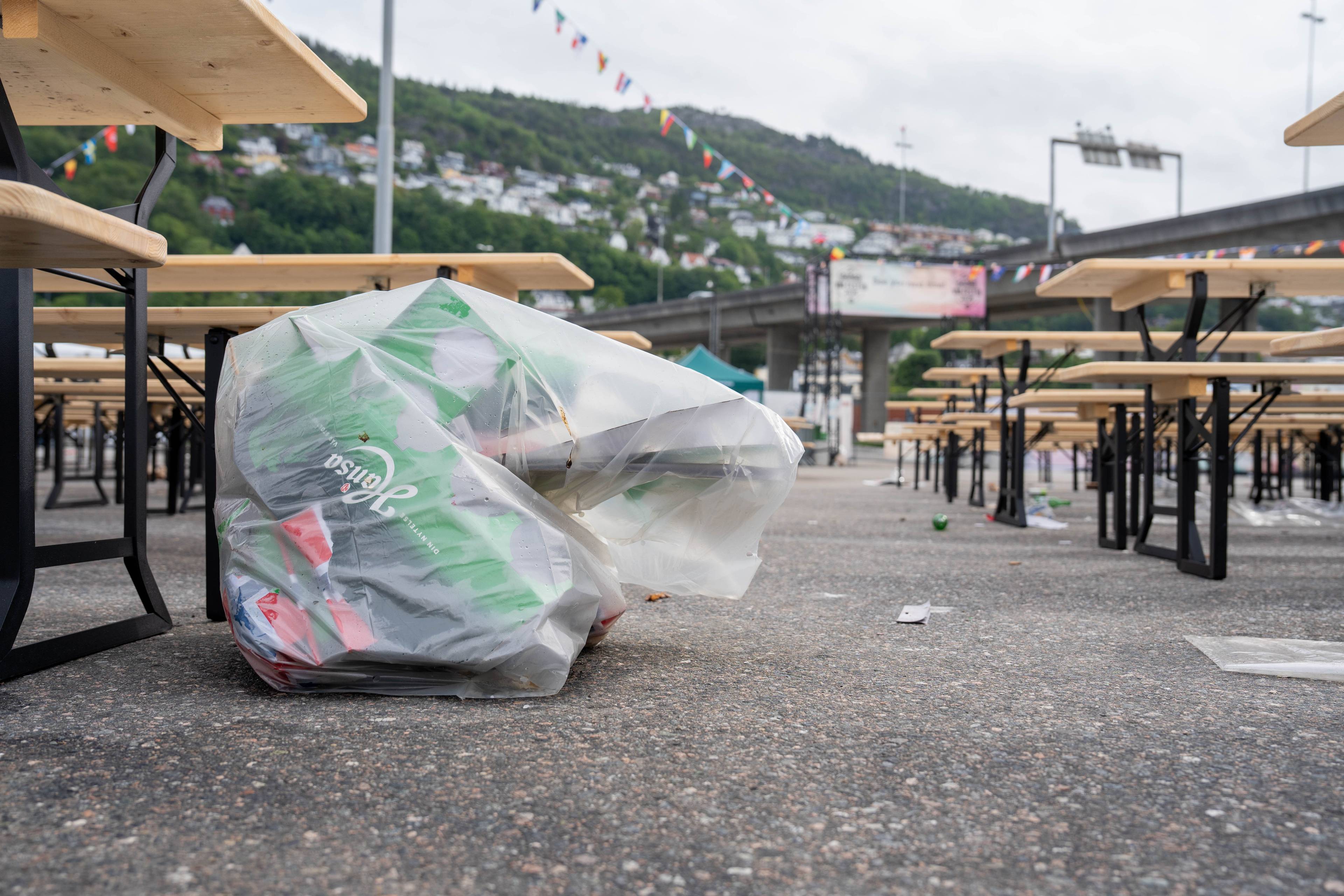 Ein søppelpose med panteflasker ligg på bakken ved fleire benker og bord. Bildet viser ein utandørs arena med ein bakke busetnad og bru i bakgrunnen. Det er ein del søppel spreidd rundt på bakken. (Bildebeskrivelsen er laget av ein KI-teneste)