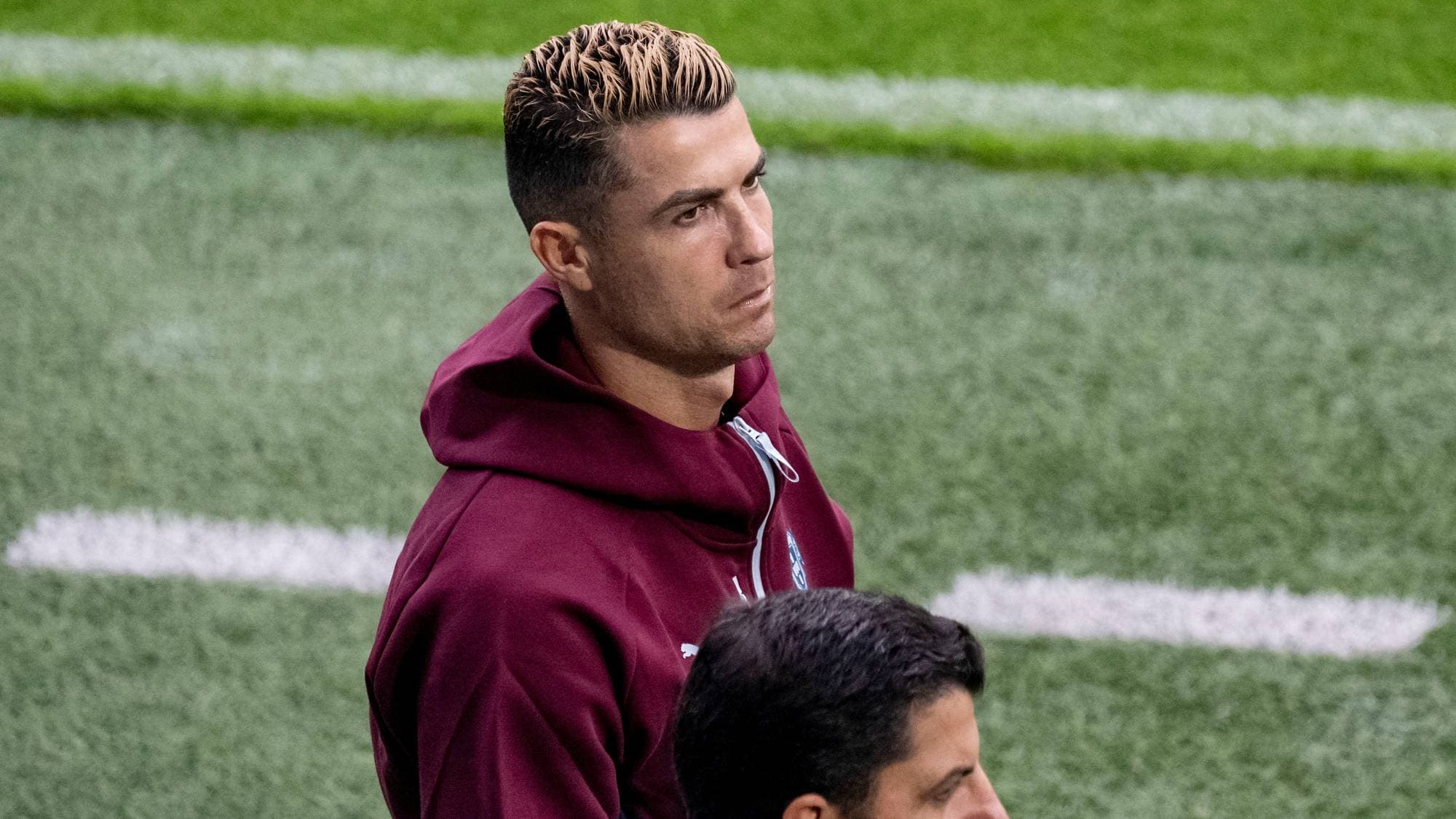 Cristiano Ronaldo i vinrød jakke på en fotballbane. En annen person sees delvis i forgrunnen. (Bildebeskrivelsen er laget av en KI-tjeneste)