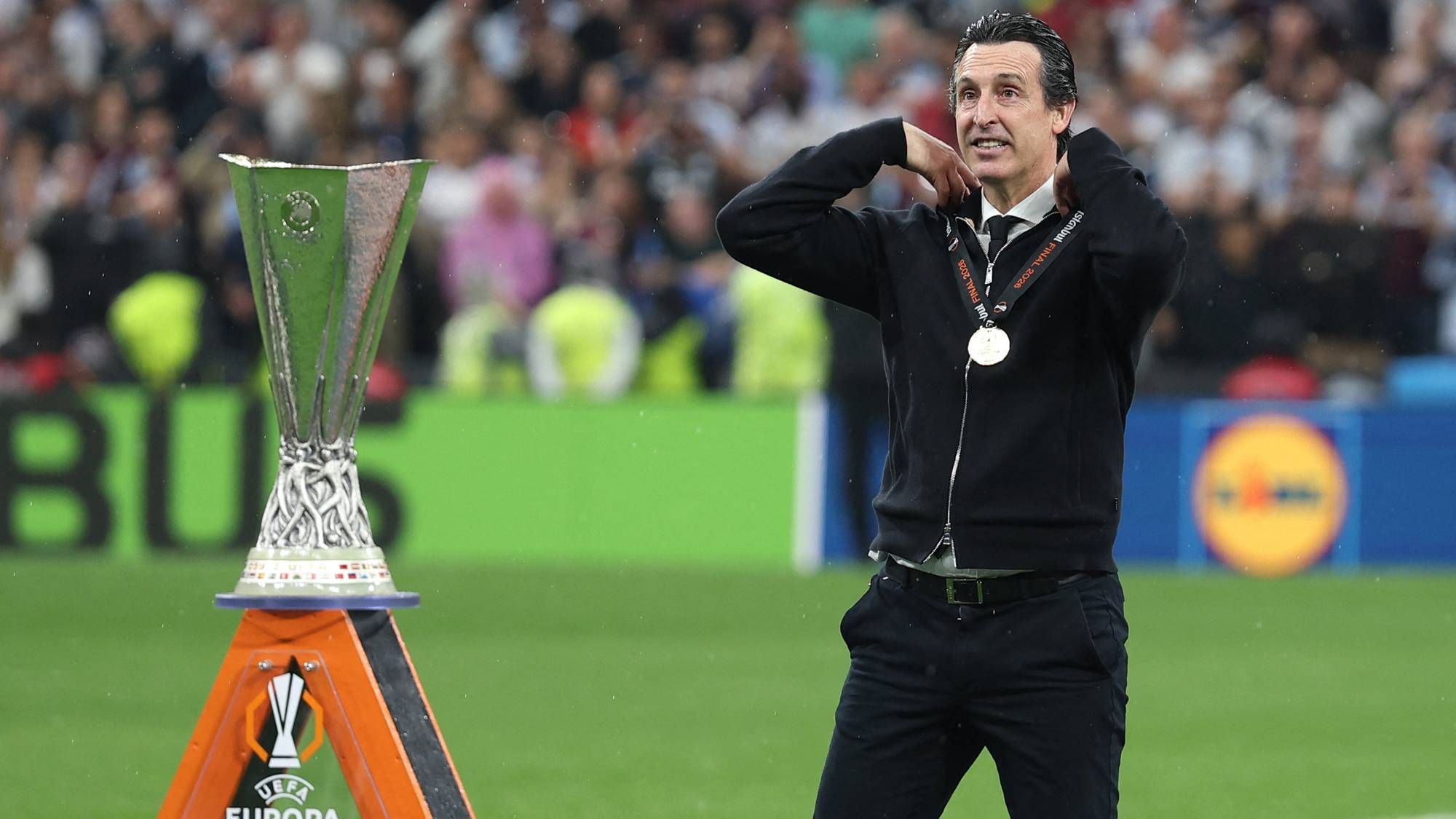 Treneren Unai Emery står ved siden av Europaliga-trofeet. Han har medalje rundt halsen. Bildet er tatt på en fotballbane med publikum i bakgrunnen. (Bildebeskrivelsen er laget av en KI-tjeneste)