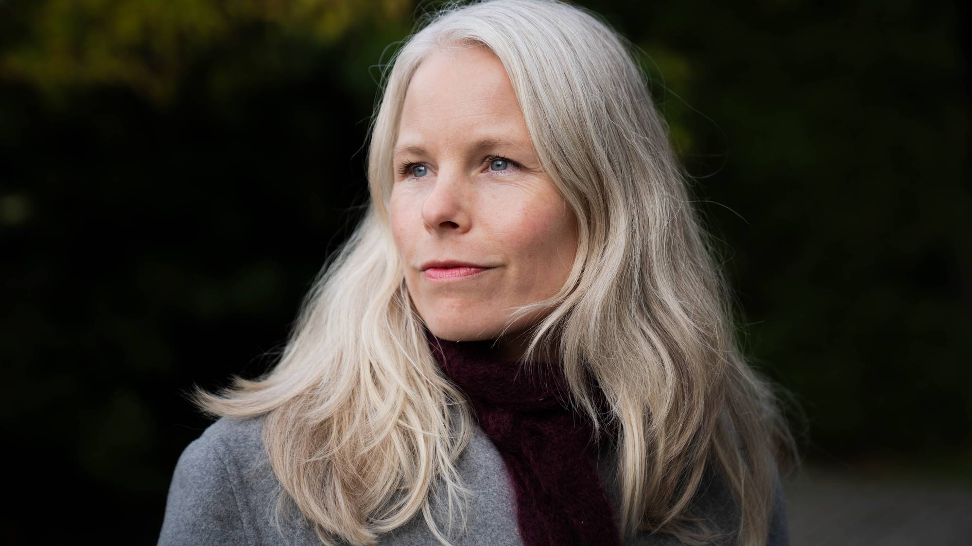 Kirsti Bergstø ser ut til høyre. Hun har lyst langt hår, høyhalset svart genser og en grå jakke. Bakgrunnen er mørk, men det skimtes at hun er ute fordi det synes noen blader. 