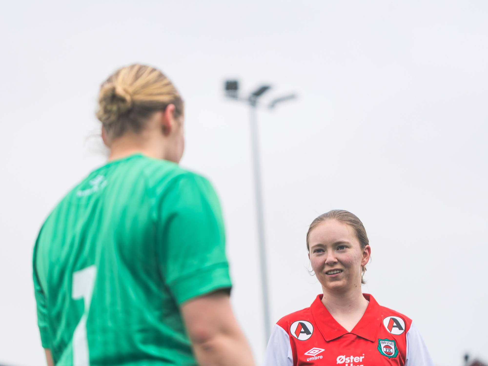 Karina står ute og snakker med sin lagkamerat. Det ser kaldt ut. Vi ser en stor lyskaster som man ofte ser på sportsarenaer i bakgrunnen. Karina har på seg rød fotballdrakt. Lagkameraten har på seg grønn, hun er keeper.