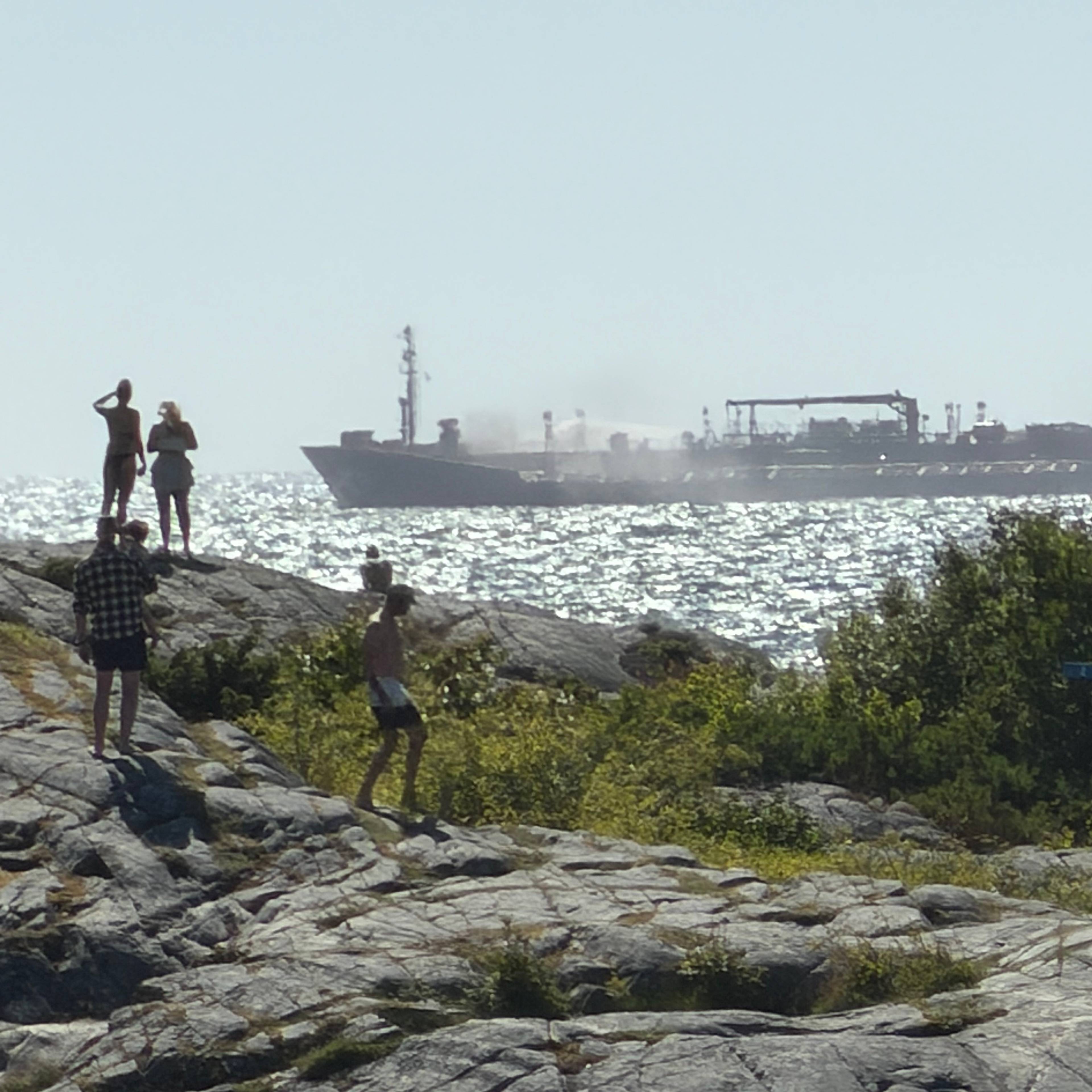 Folk står på svaberg og ser ut mot et lasteskip med røykutvikling på havet. (Bildebeskrivelsen er laget av en KI-tjeneste)