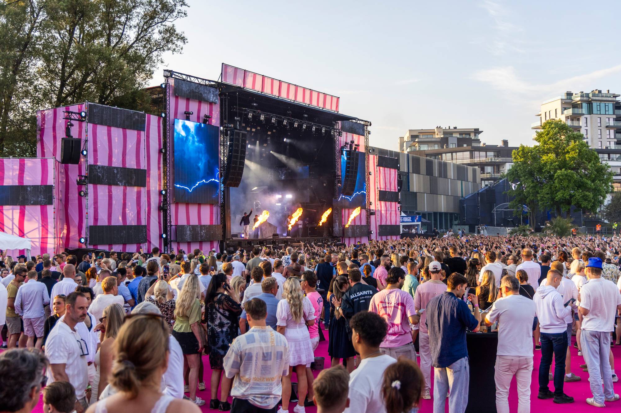 Stort publikum foran en festivalscene med pyrotekniske flammer og vertikale rosa striper. (Bildebeskrivelsen er laget av en KI-tjeneste)
