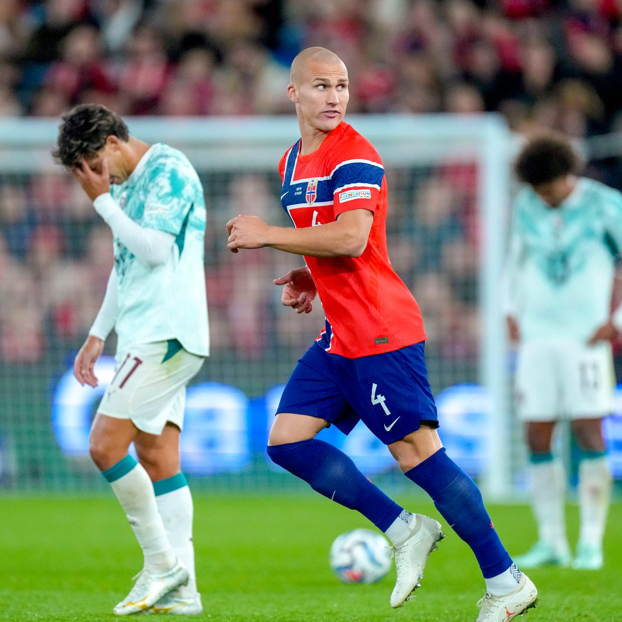 Norsk fotballspiller i rød trøye løper på banen med motstandere i bakgrunnen. (Bildebeskrivelsen er laget av en KI-tjeneste)