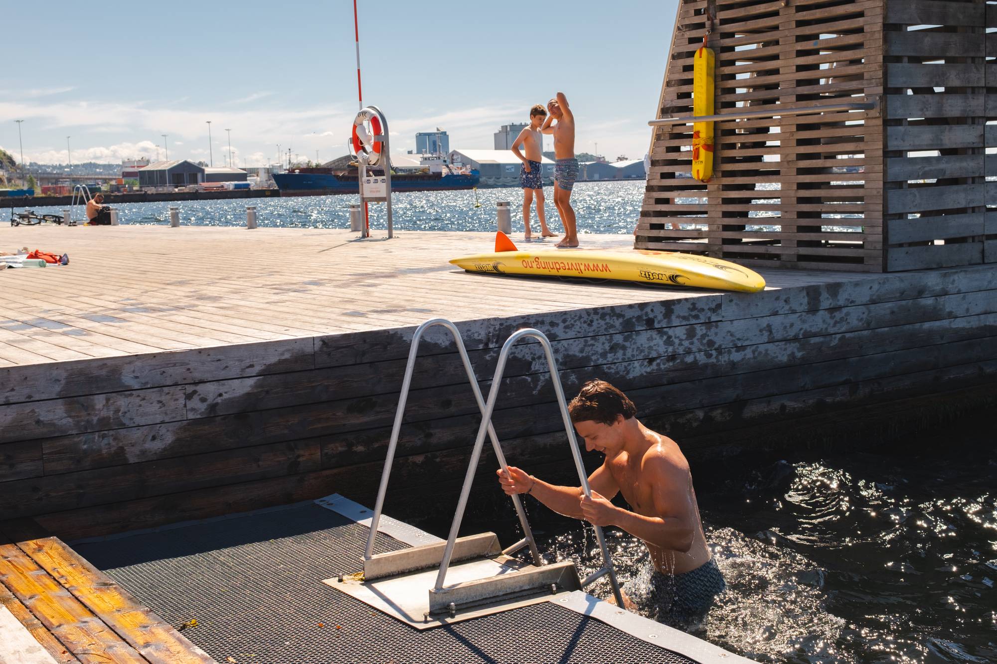 Personer bader fra en trebrygge på Sørenga Sjøbad i Oslo på en solskinnsdag. (Bildebeskrivelsen er laget av en KI-tjeneste)