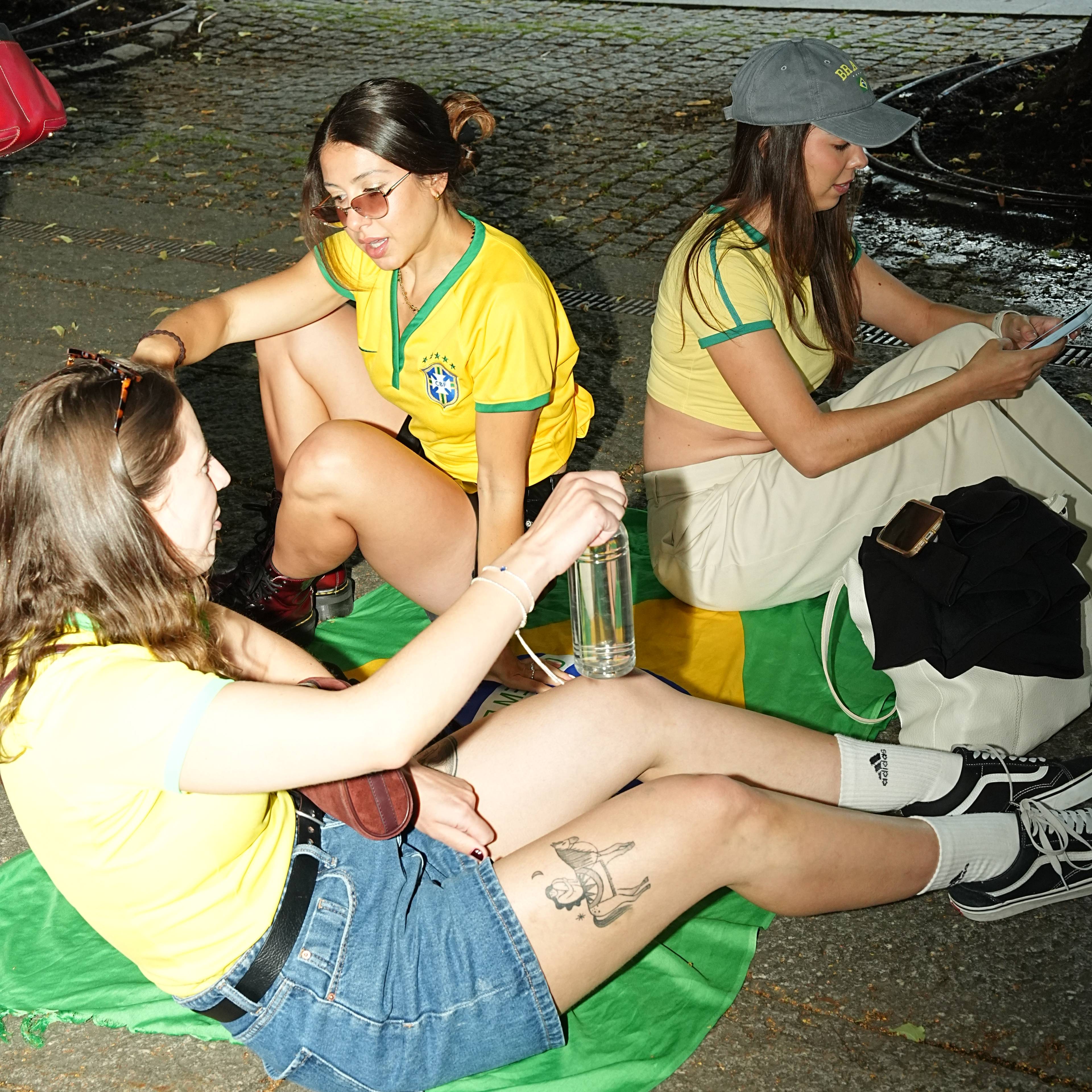 Tre kvinner sitter på et brasiliansk flagg på en gate mens de bruker mobiltelefoner. (Bildebeskrivelsen er laget av en KI-tjeneste)