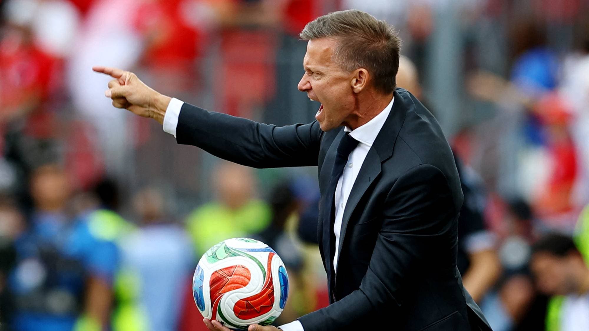 En mann i dress holder en fotball og peker med den ene hånden. Han er på en fotballbane med tilskuere i bakgrunnen. (Bildebeskrivelsen er laget av en KI-tjeneste)
