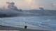 Foto: Aziz Taher / Reuters To personer går bortover en strand i morgentimene. Det blå havet topper seg i hvitt når det slår i bølger innover mot den blekgrå sanden. I bakgrunnen velter grålilla røyk opp fra en by etter rakettangrep fra Israel inn i det sørlige Libanon.