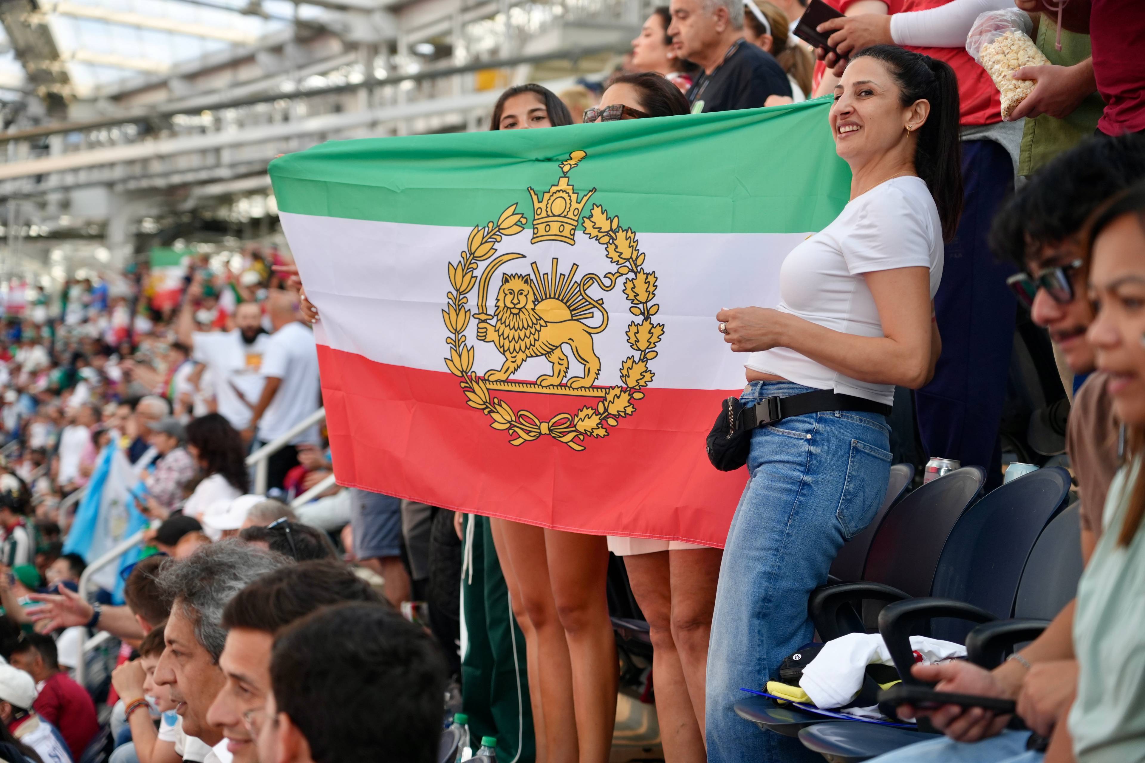 En kvinne holder et iransk flagg foran en mengde på et stadion. Flagget har grønn, hvit og rød farge. I midten er et våpenskjold med en løve. Flere personer ses i bakgrunnen og i forgrunnen. (Bildebeskrivelsen er laget av en KI-tjeneste)