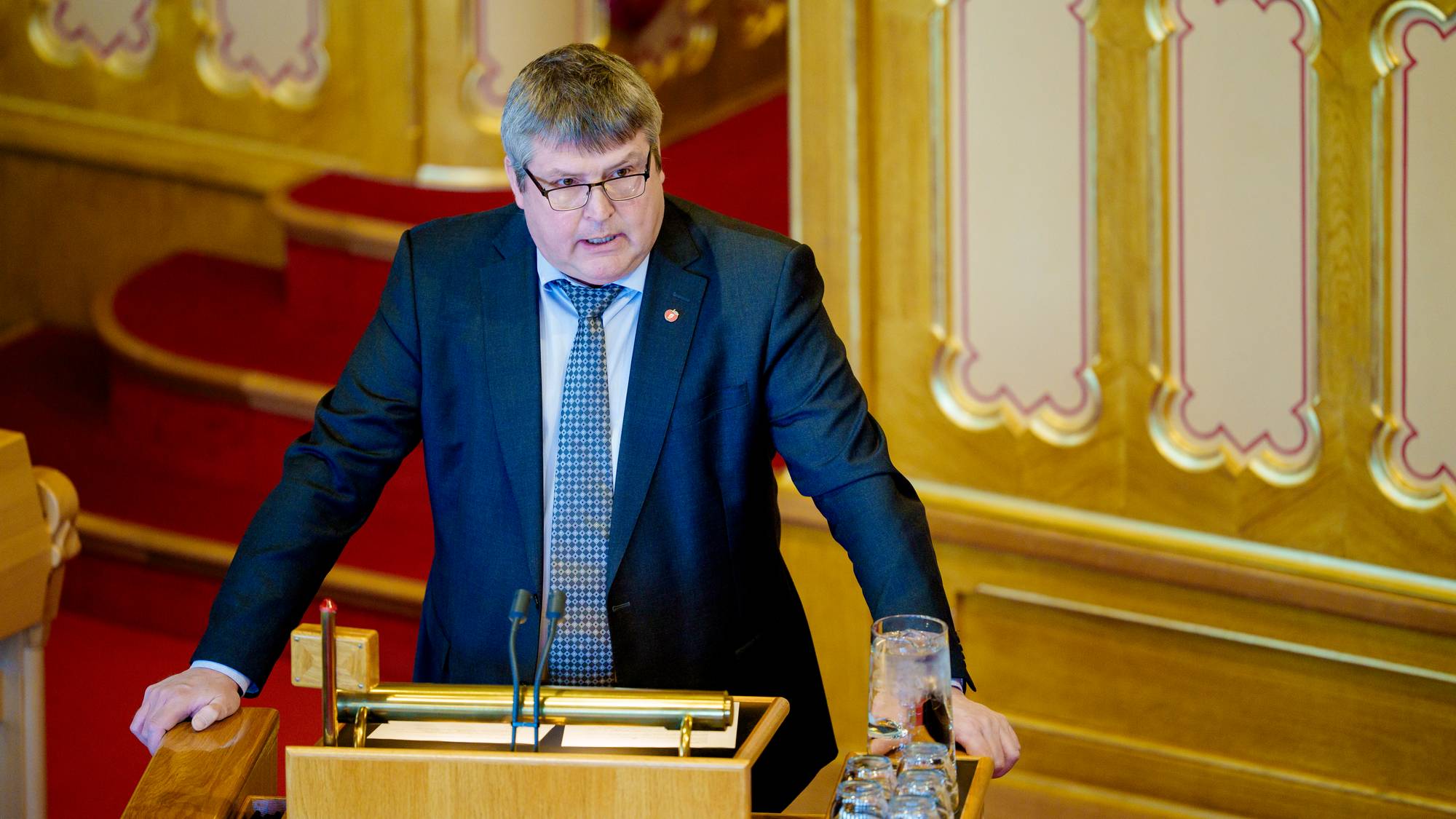 En mann i dress med briller taler foran Stortingets talerstol.