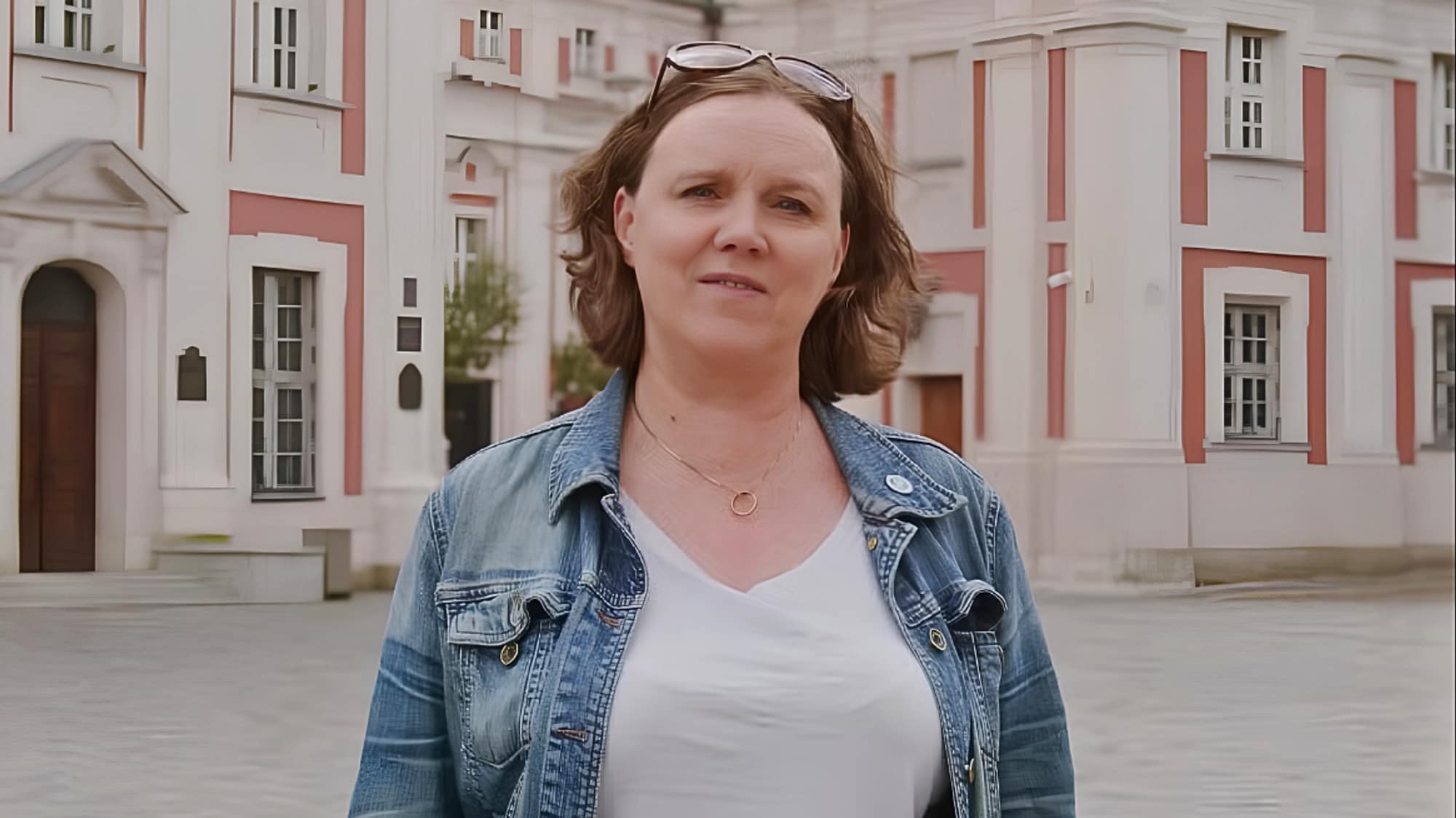 Kvinne med briller på hodet foran en eldre bygning med hvite og røde detaljer. 