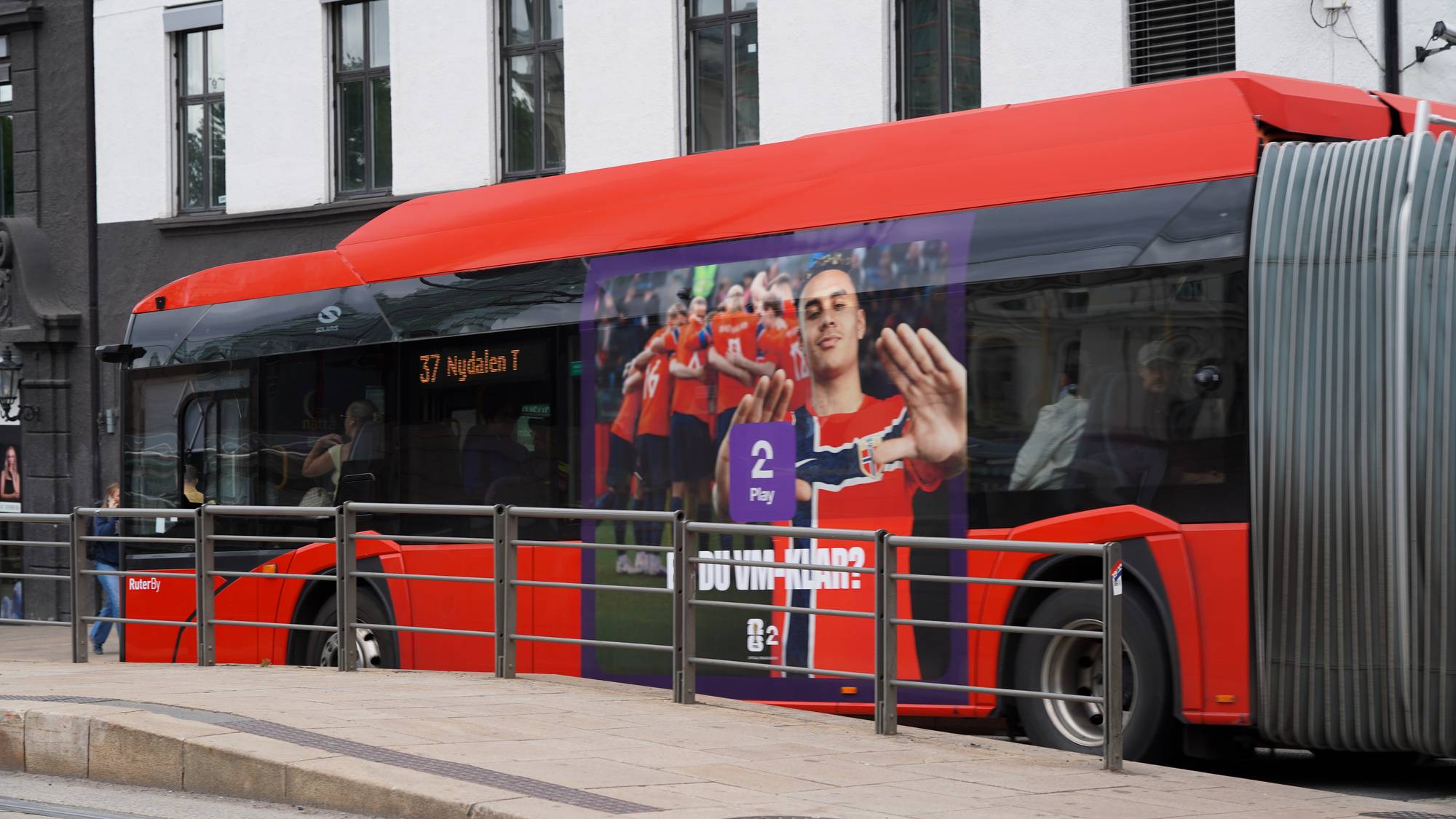 En rød buss med reklame kjører forbi en bygning. På reklamen vises et bilde av fotballspillere på det norske herrelandslaget. Bussen har linje nummer 37 og destinasjon Nydalen T. (Bildebeskrivelsen er laget av en KI-tjeneste)