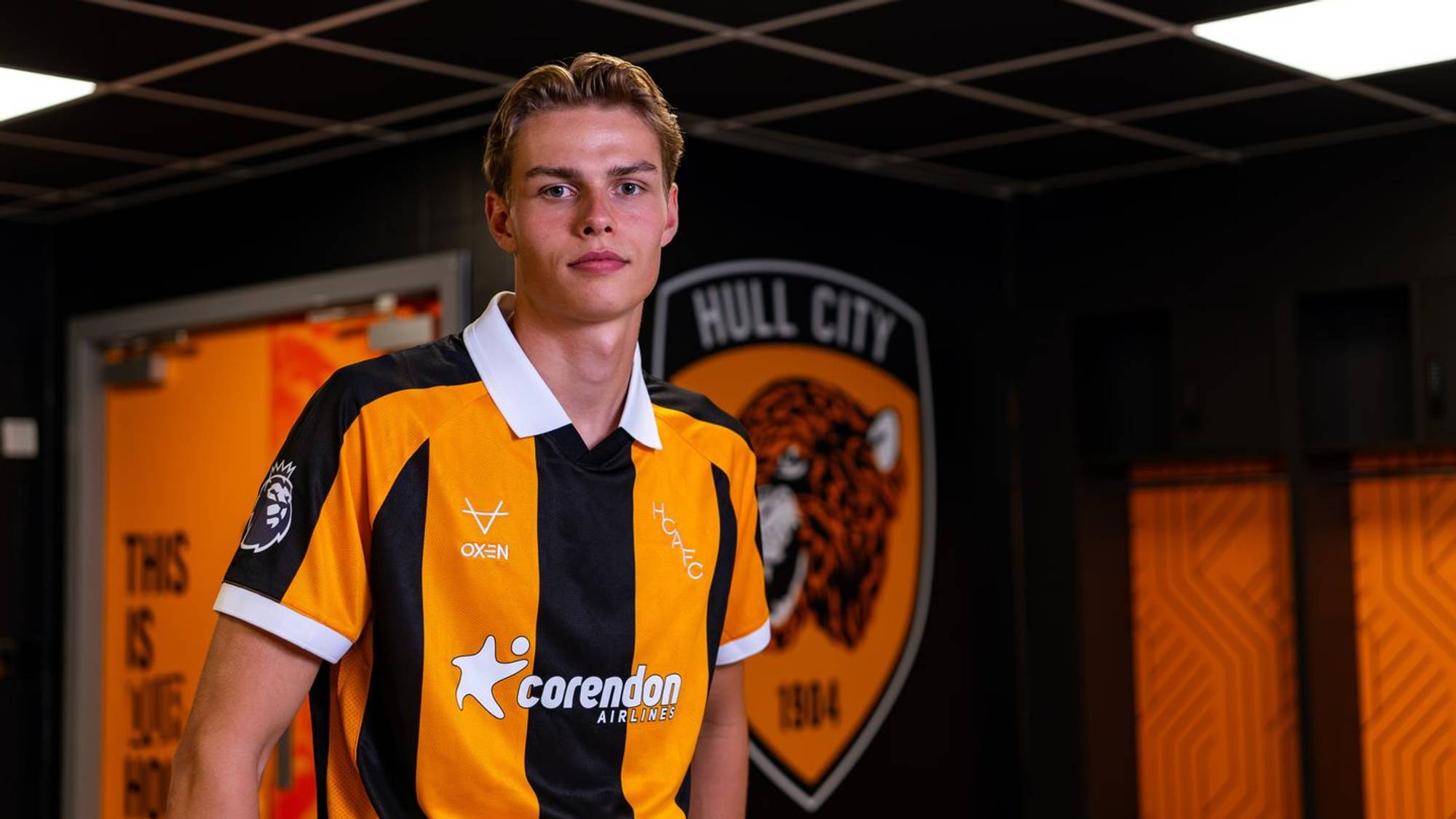 Fotballspiller i Hull City-drakt står i et garderobeområde med klubbens logo i bakgrunnen. (Bildebeskrivelsen er laget av en KI-tjeneste)