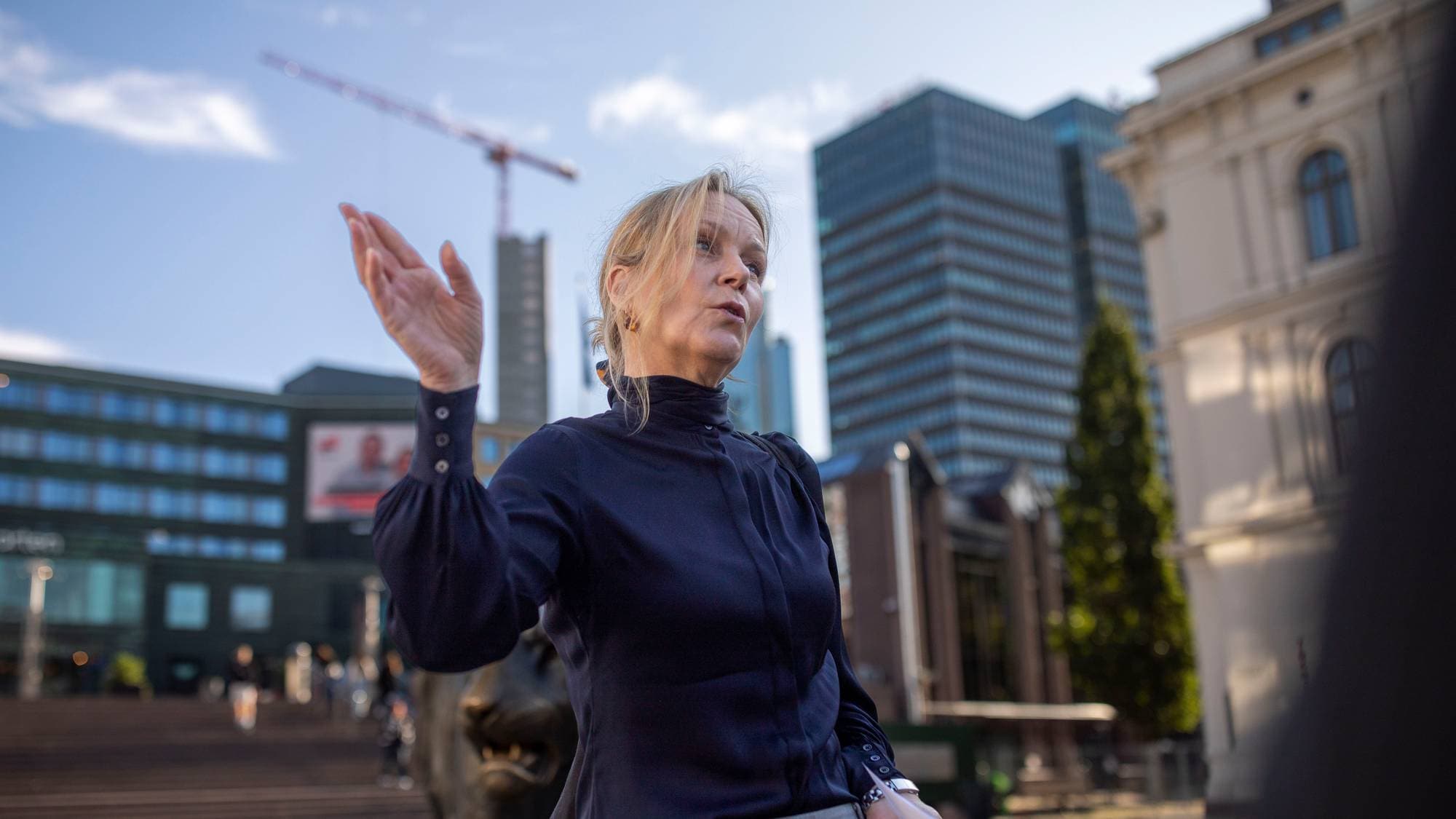 Kvinne i mørk bluse snakker og gestikulerer utendørs i en by med moderne kontorbygg i bakgrunnen. (Bildebeskrivelsen er laget av en KI-tjeneste)