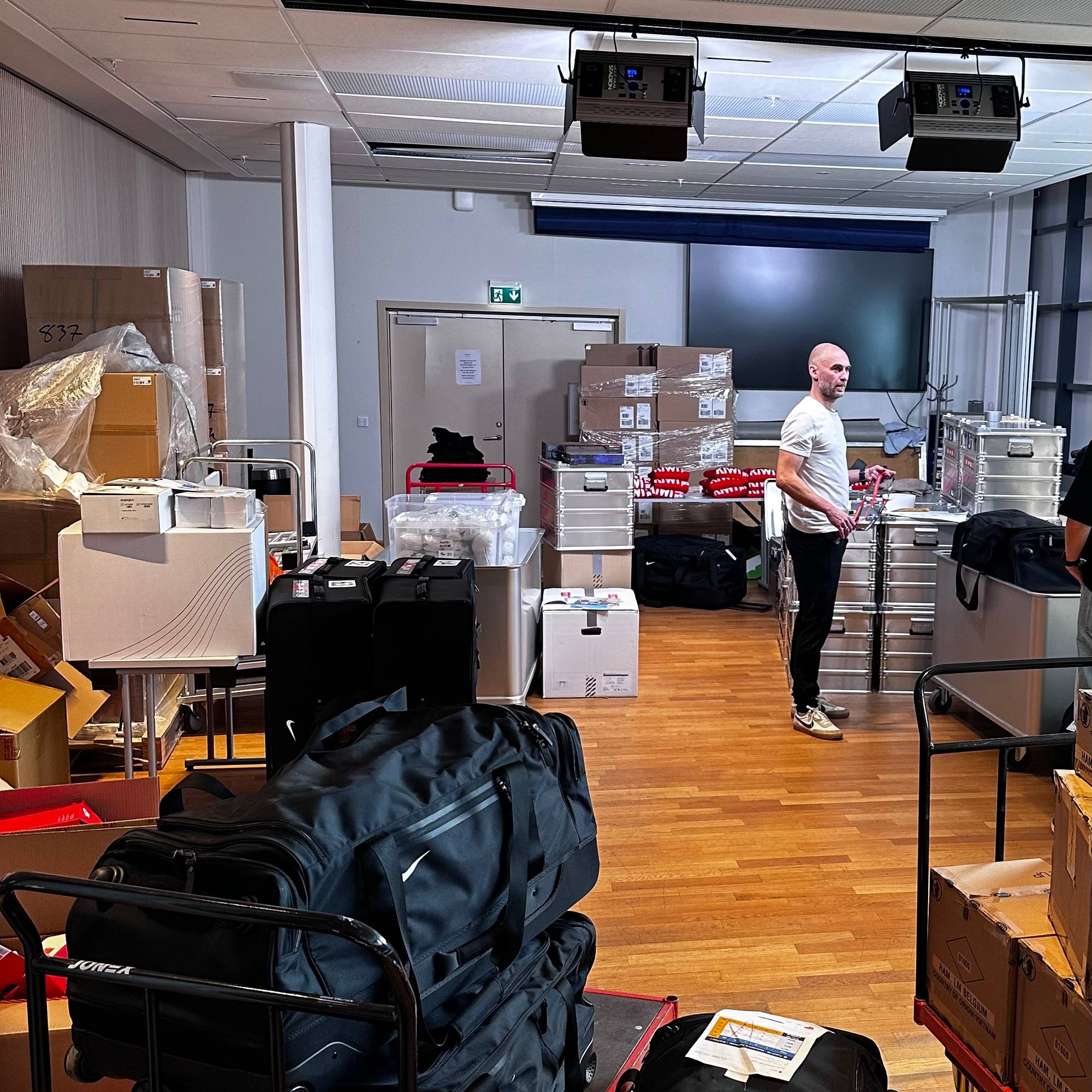 Mann står i et rom fylt med esker og bagger. Mannen er kledd i en hvit t-skjorte og mørke bukser. Rommet har tregulv og et nedforingspanel i taket. Det er flere traller med store svarte bager og stabler med esker på gulvet. (Bildebeskrivelsen er laget av en KI-tjeneste)