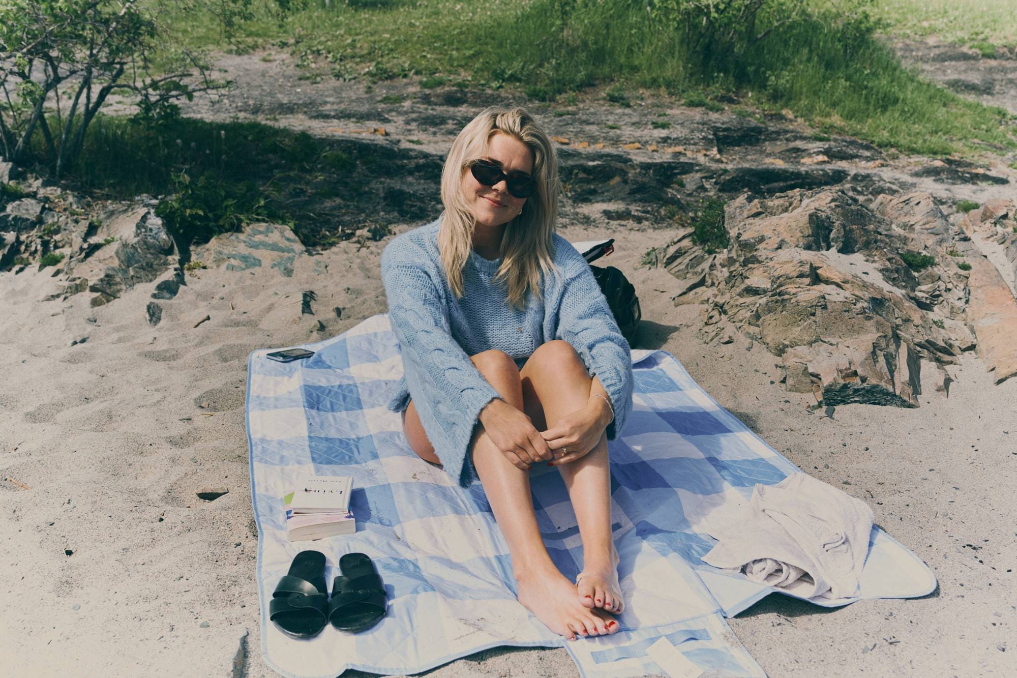 Ung kvinne med lyst hår og solbriller sitter i en genser med bare ben på et teppe på stranden. 