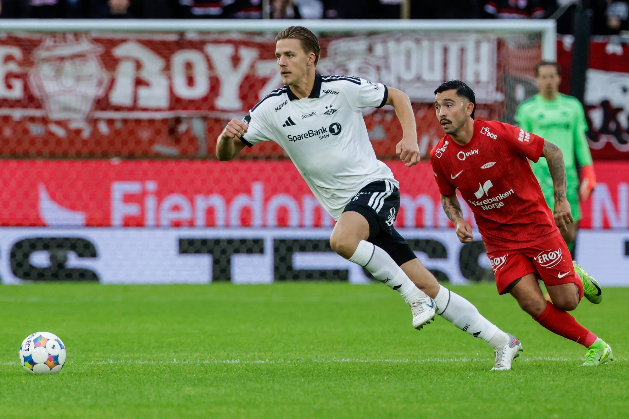 Fotballspillere fra Rosenborg og Brann løper etter ballen på en fotballbane. (Bildebeskrivelsen er laget av en KI-tjeneste)