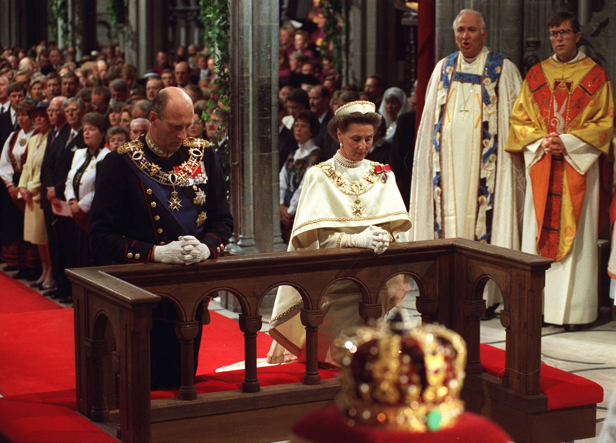 Kong Harald og dronning Sonja kneler ved alteret under signingen i Nidarosdomen i 1991. I forgrunnen står kongekronen på en rød fløyelsespute.