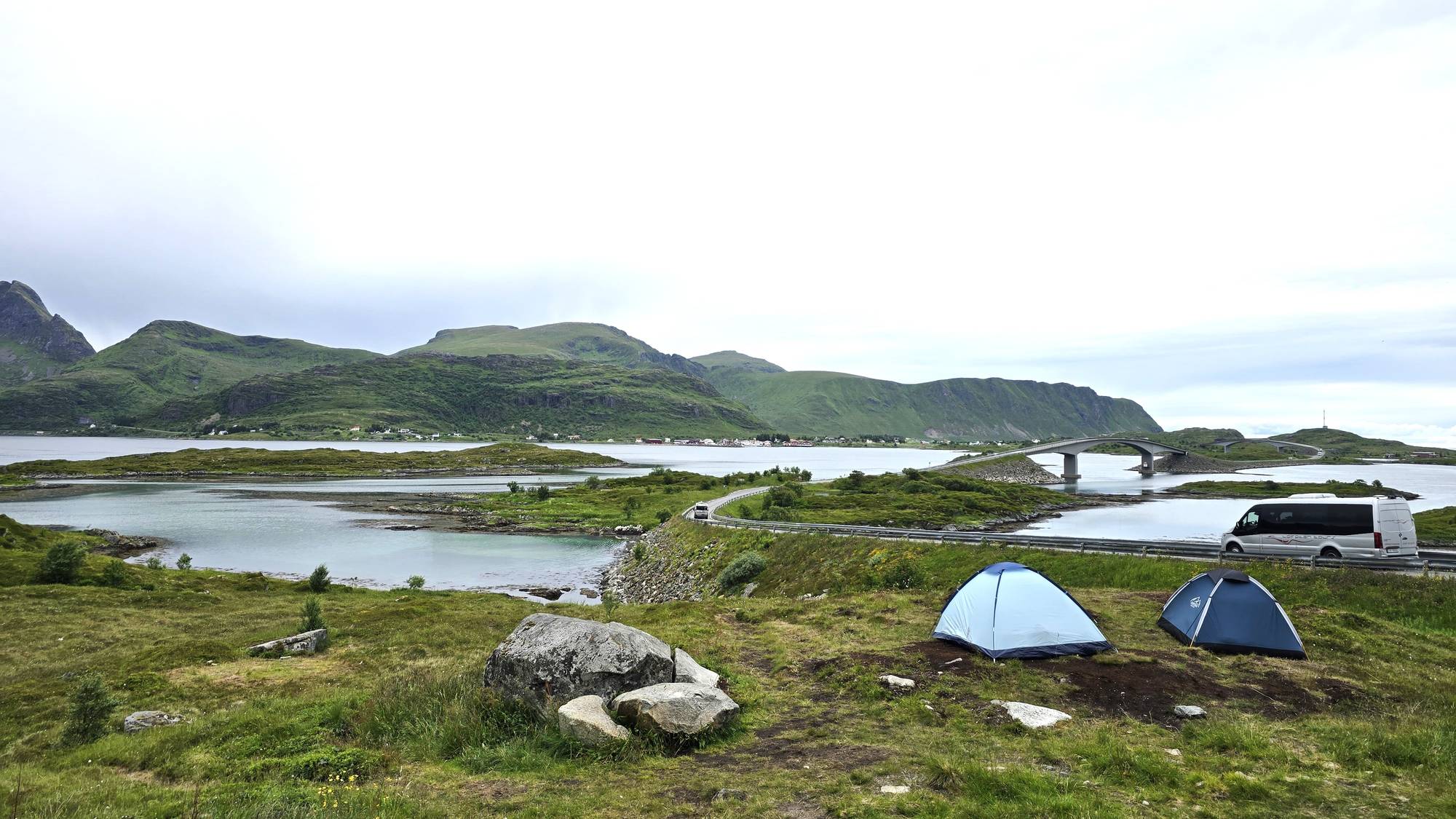 Flere turister har satt opp telt langs veien i Lofoten. Veien slynger seg over broer og odder i fjorden og man ser grønne fjell i bakgrunn.
