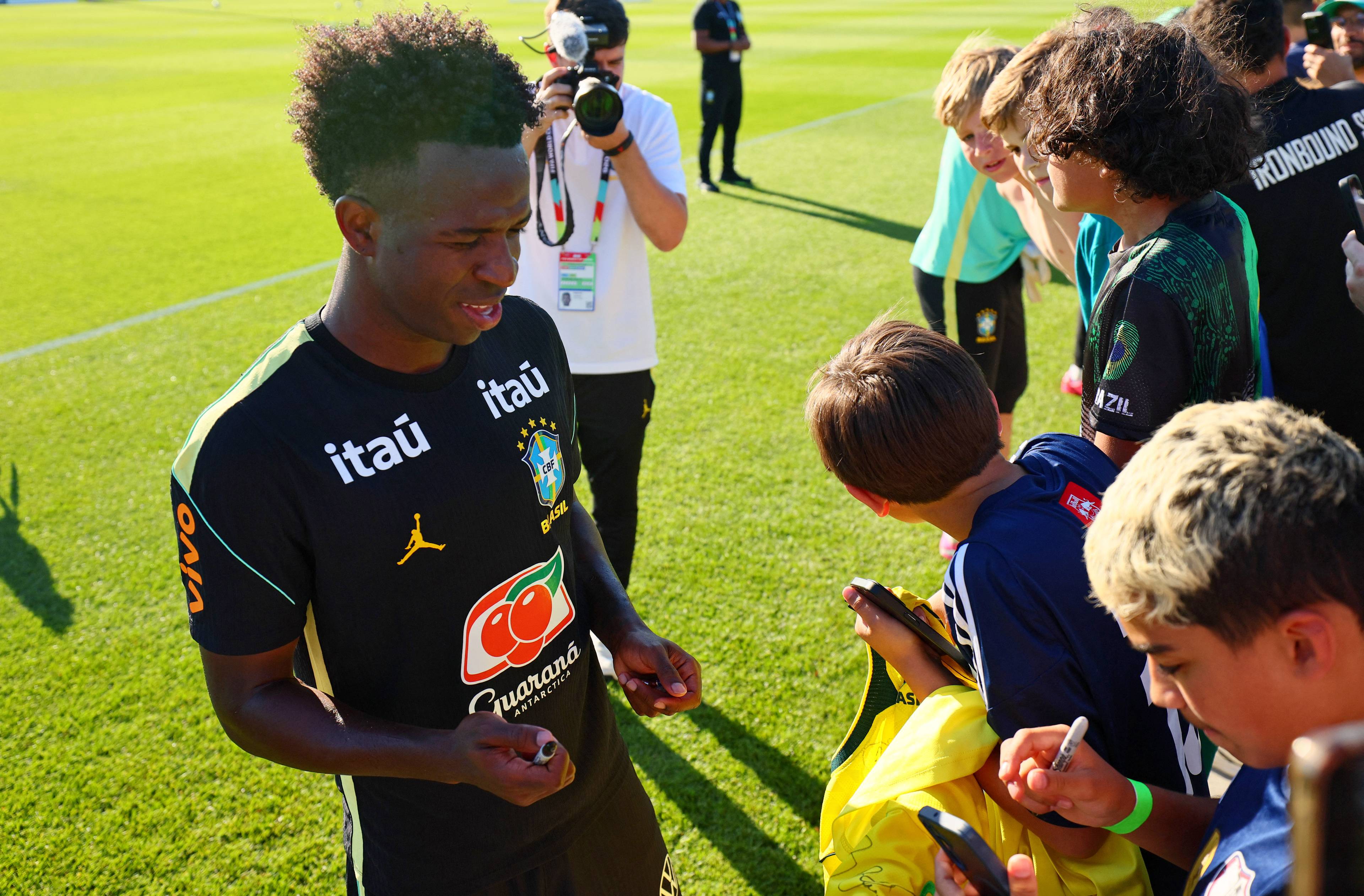 Fotballspilleren Vincius Jnior signerer autografer for unge supportere på en fotballbane. Flere barn står rundt ham og holder frem autografskriver og mobiltelefoner. En fotograf tar bilde i bakgrunnen. (Bildebeskrivelsen er laget av en KI-tjeneste)