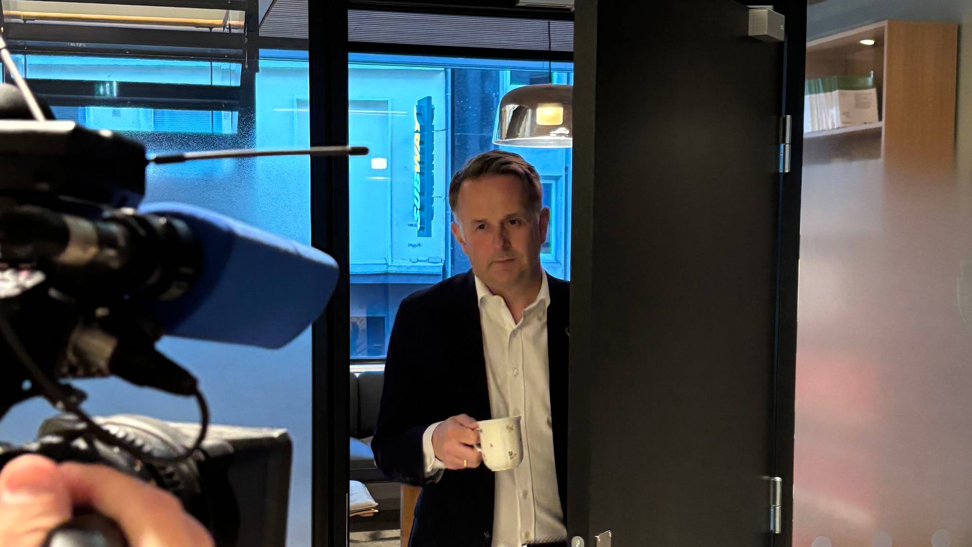 En mann i dress går ut gjennom en dør. Et TV-kamera venter på ham.