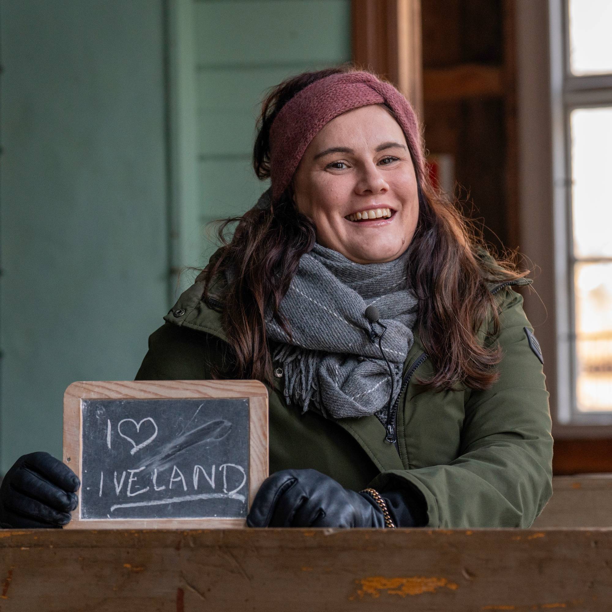 En kvinne med pannebånd og jakke smiler og holder opp et skilt der det står "I love Iveland"
