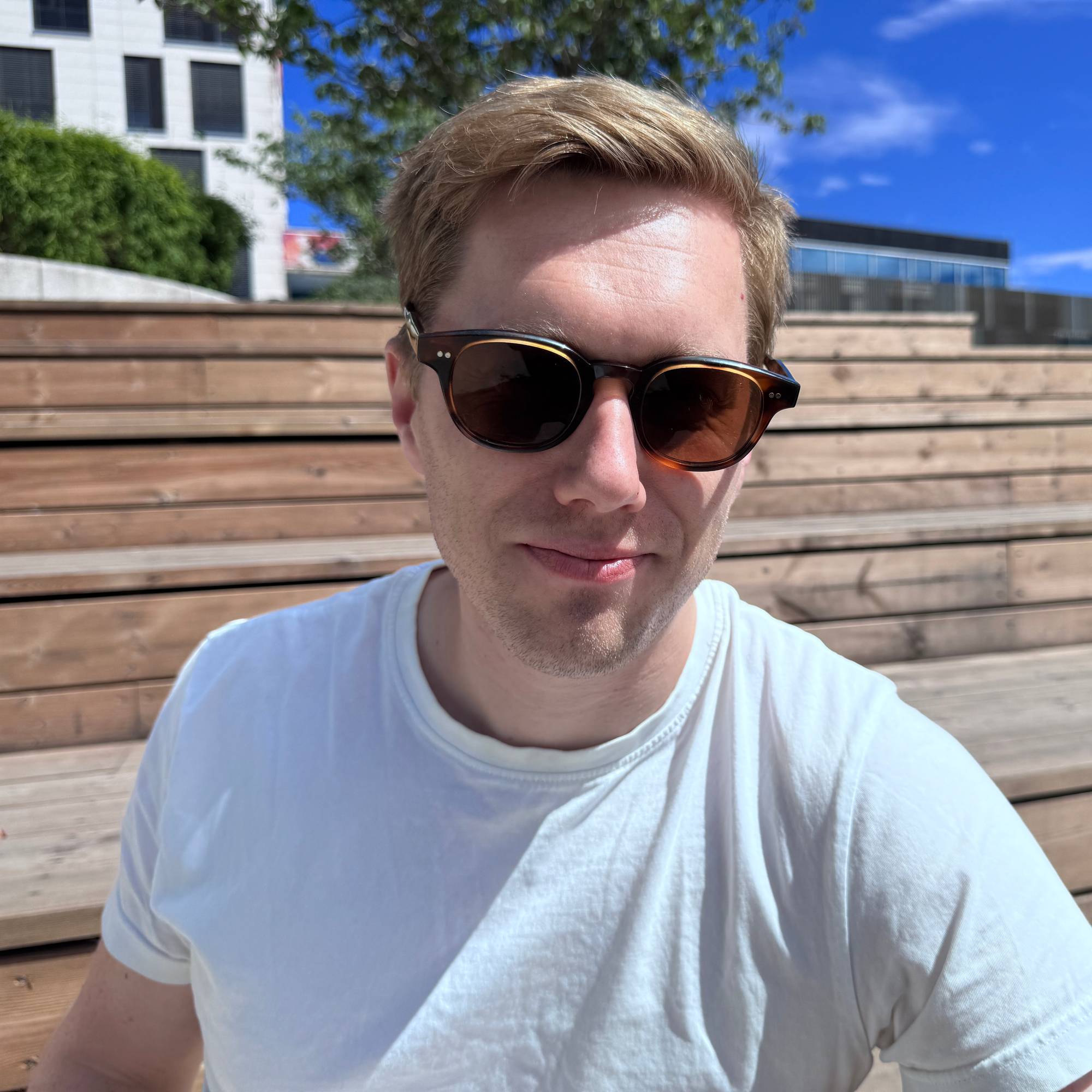 En mann med solbriller og hvit t-skjorte sitter i en trapp. I bakgrunnen ser man en bygning.