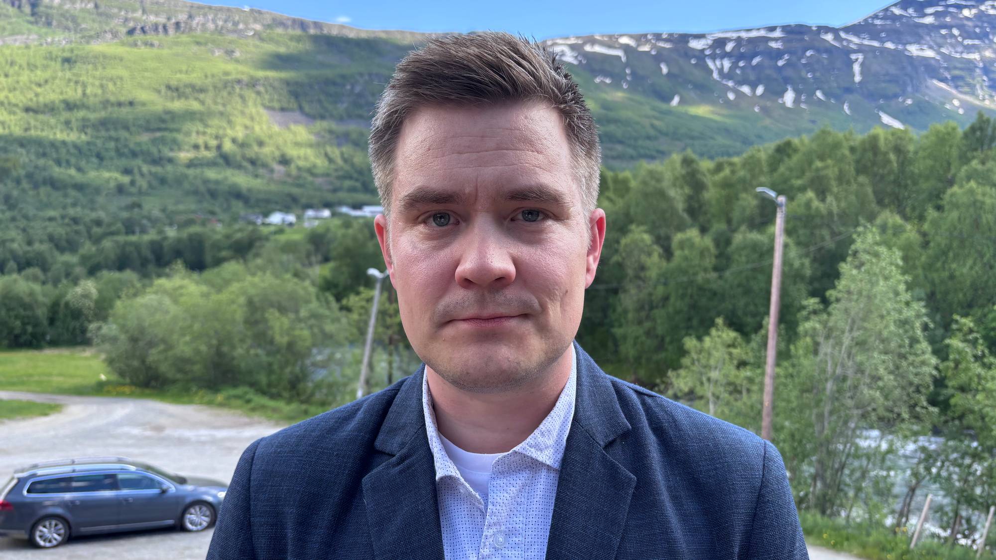 En mann i dress ser i kamera. En bil og natur i bakgrunnen, man skimter en elv.