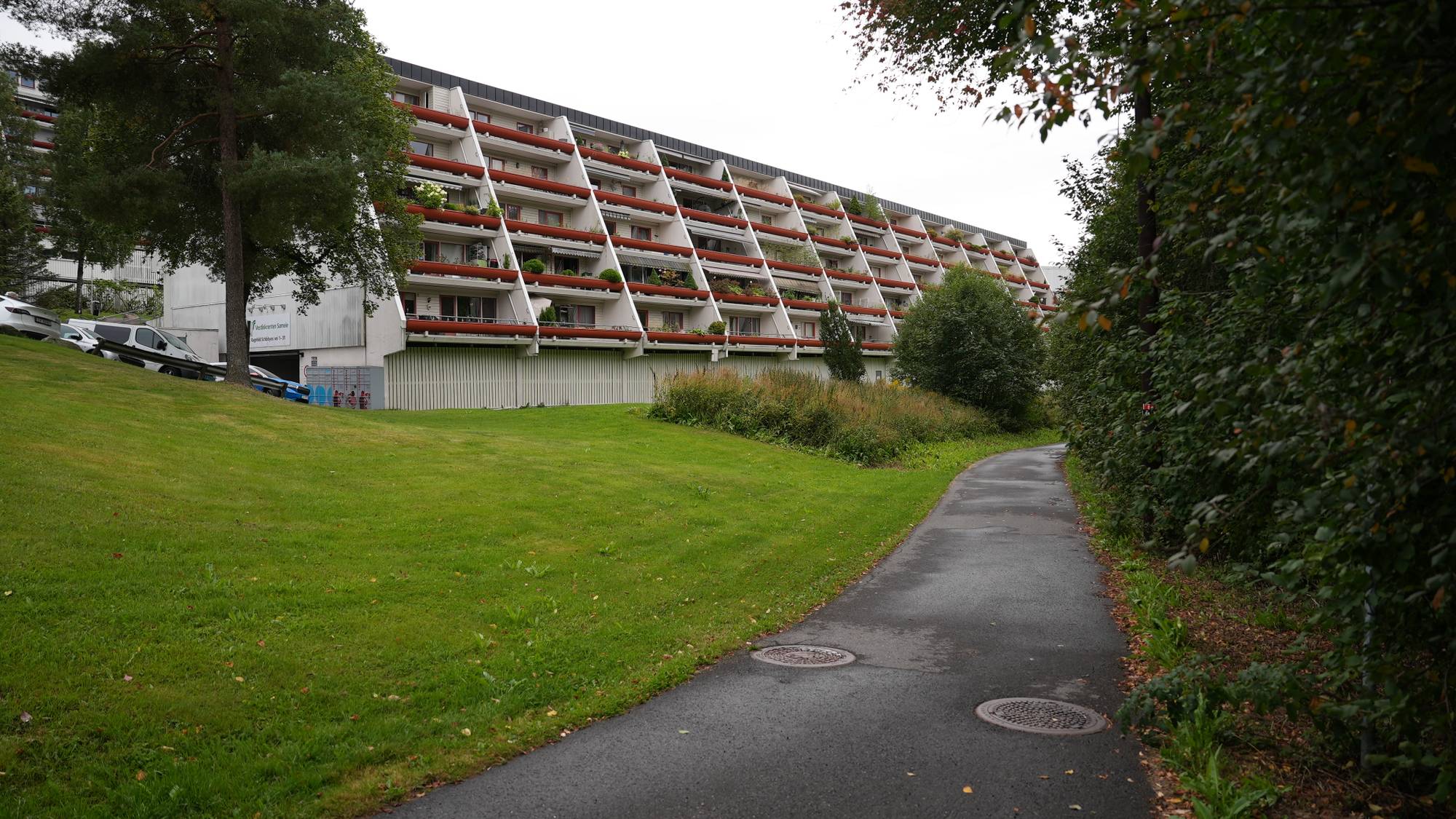 En lang blokk med terrasser sett fra en gangsti med grøntarealer. (Bildebeskrivelsen er laget av en KI-tjeneste)