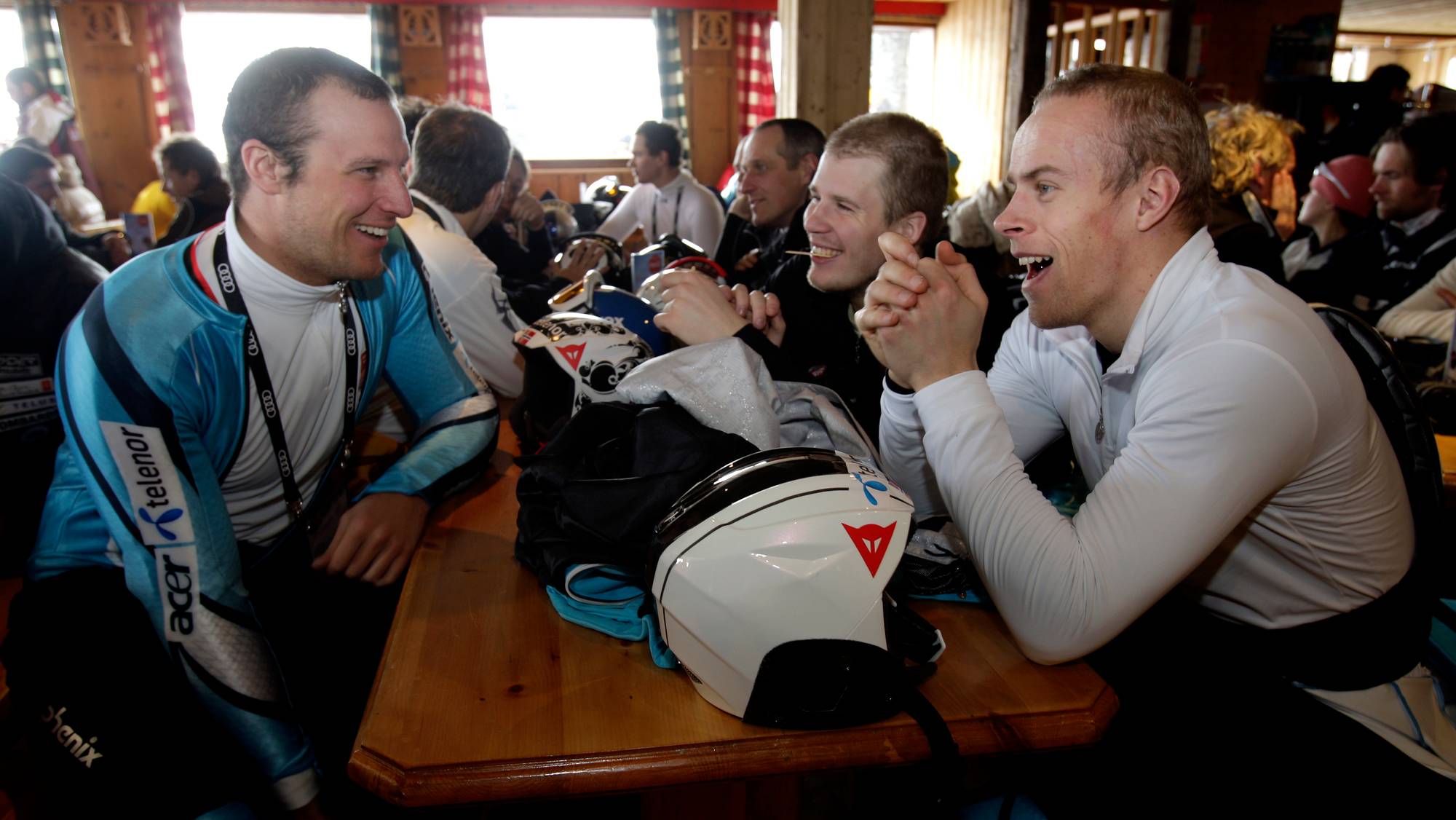 Menn i skibukser og dresser sitter ved et bord med hjelmer. Mennene ser ut til å prate og smile til hverandre. Bakgrunnen er en innendørs setting med trevegger og vinduer. (Bildebeskrivelsen er laget av en KI-tjeneste)