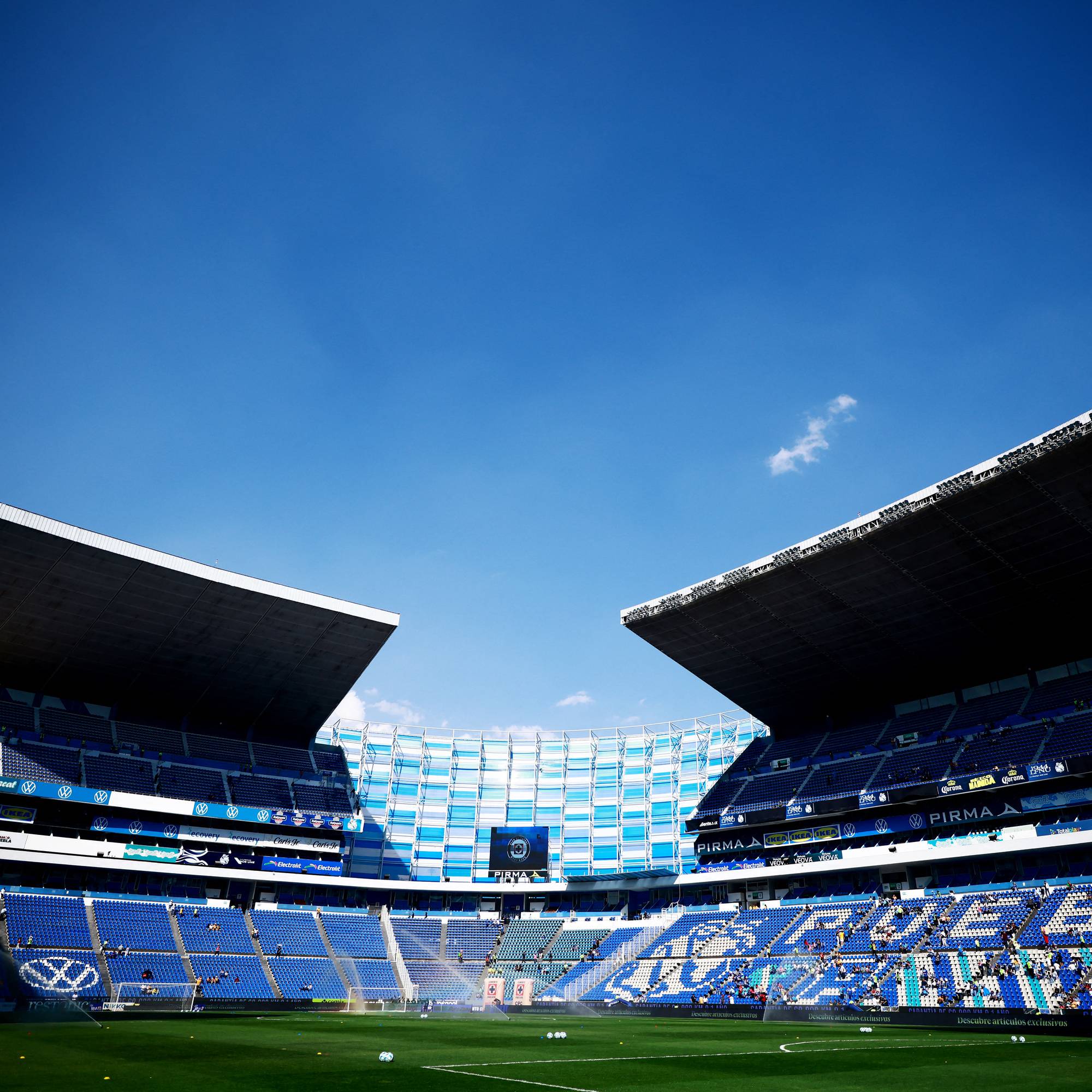 Et stort fotballstadion med blå tribuner og et grønt gressfelt er vist under en klar blå himmel. Mange seter på tribunene er tomme, mens noen få tilskuere sitter spredt. Stadionets tak har en markant, kantete design. Tekst og logoer er synlige på tribunene og på reklameskilt langs banen. (Bildebeskrivelsen er laget av en KI-tjeneste)