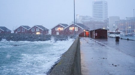 Uvær i Bodø i Nordland. Bølgene slår inn mot land over moloen i Bodø og skyller over røde sjøhus. I bakgrunn synes byen i snøvær.
