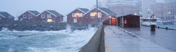 Uvêr i Bodø i Nordland. Bølgene slår inn mot land over moloen i Bodø og skyl over røde sjøhus. I bakgrunn synest byen i snøvêr.