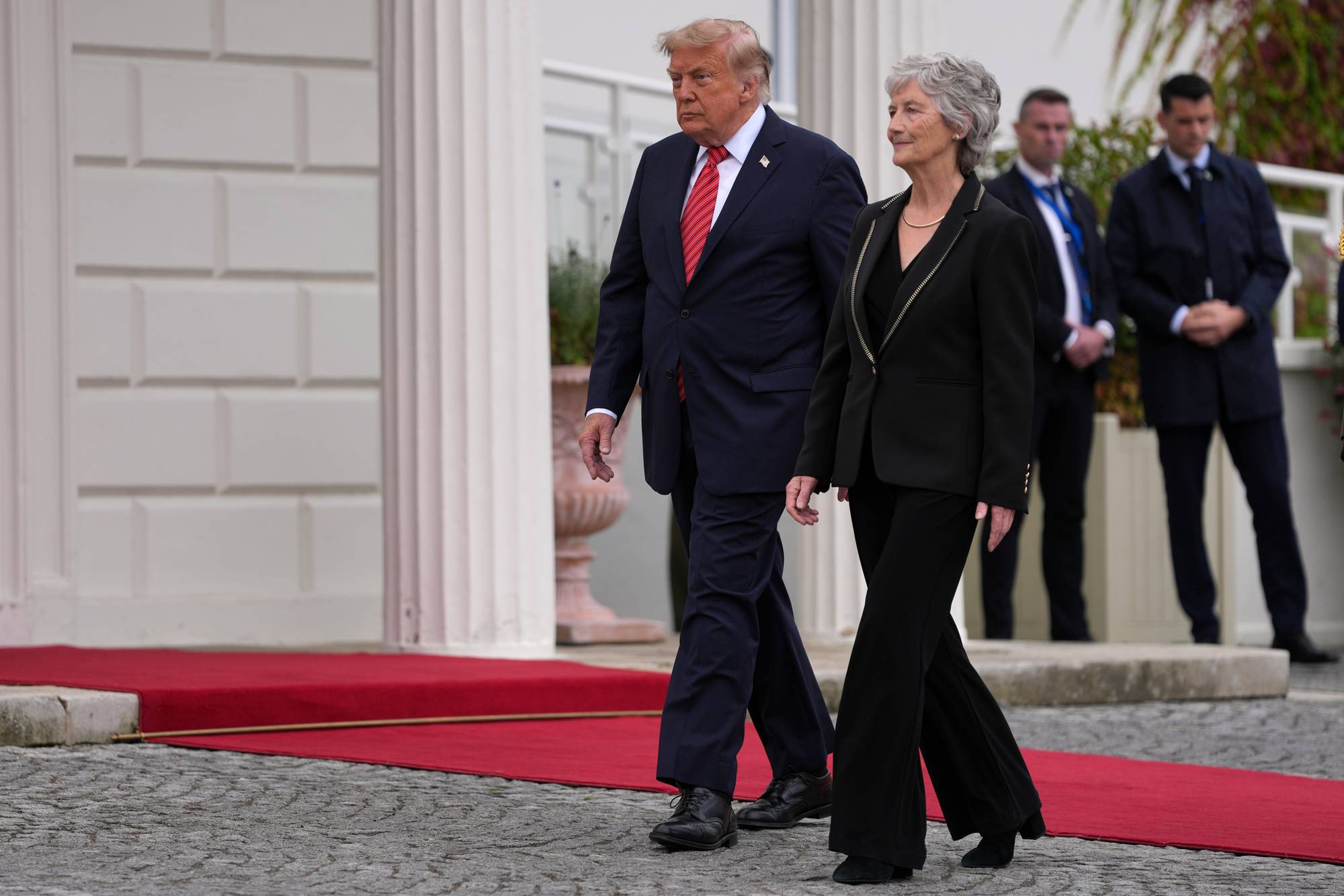 Donald Trump går sammen med en kvinne på en rød løper utenfor en bygning. Vakter står i bakgrunnen. (Bildebeskrivelsen er laget av en KI-tjeneste)