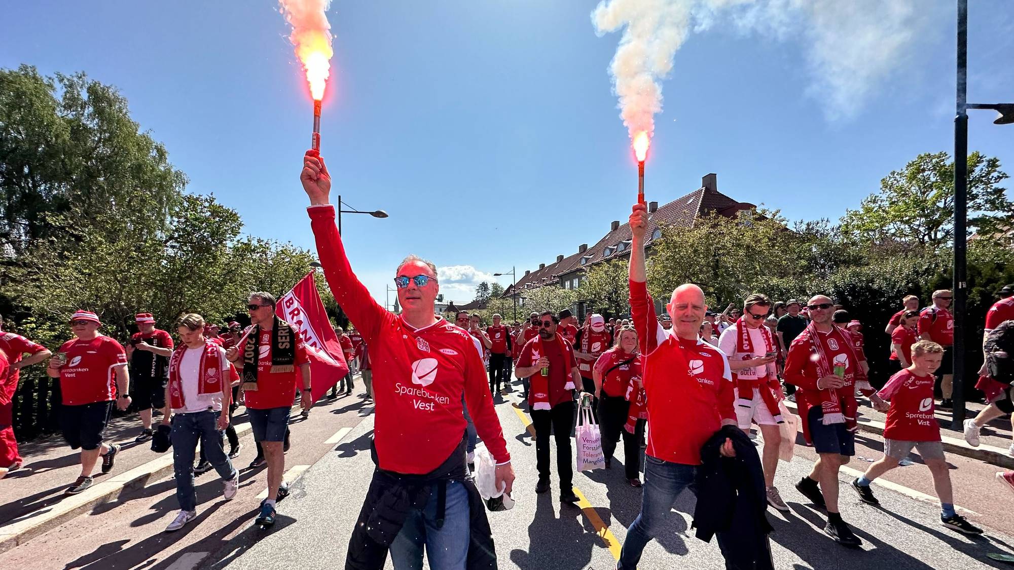 Brann-supportere kledd i røde drakter går i tog gjennom en gate. De to fremste, to skallede menn, holder fakler som lager flammer og røyk. Ved siden av veien er det grønne trær, og man ser også hustak i bakgrunnen. 