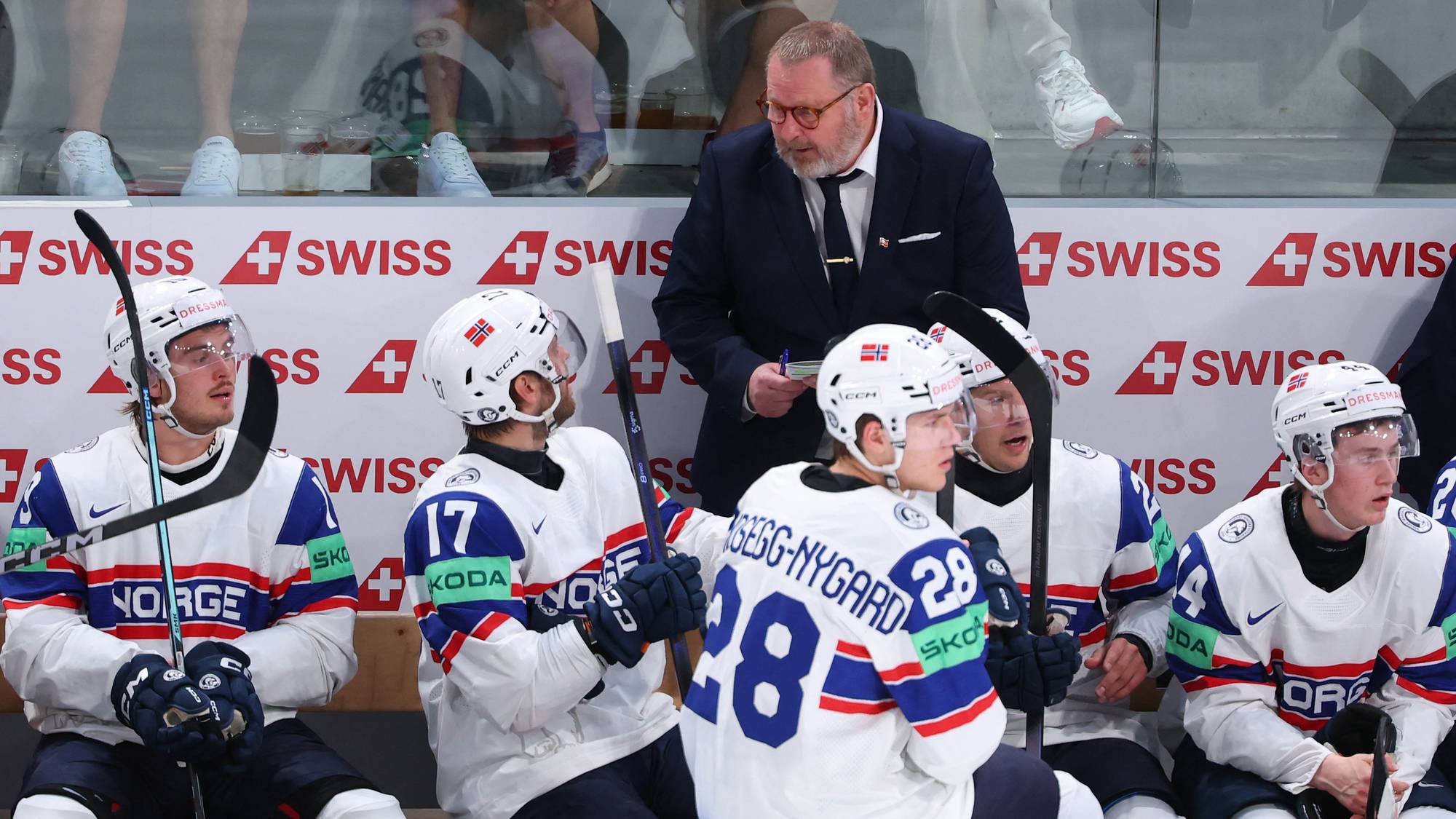 Tre ishockeyspillere i hvite drakter sitter på benken. En trener i dress står bak dem. Bak dem er det en reklamevegg med SWISS-logo. (Bildebeskrivelsen er laget av en KI-tjeneste)
