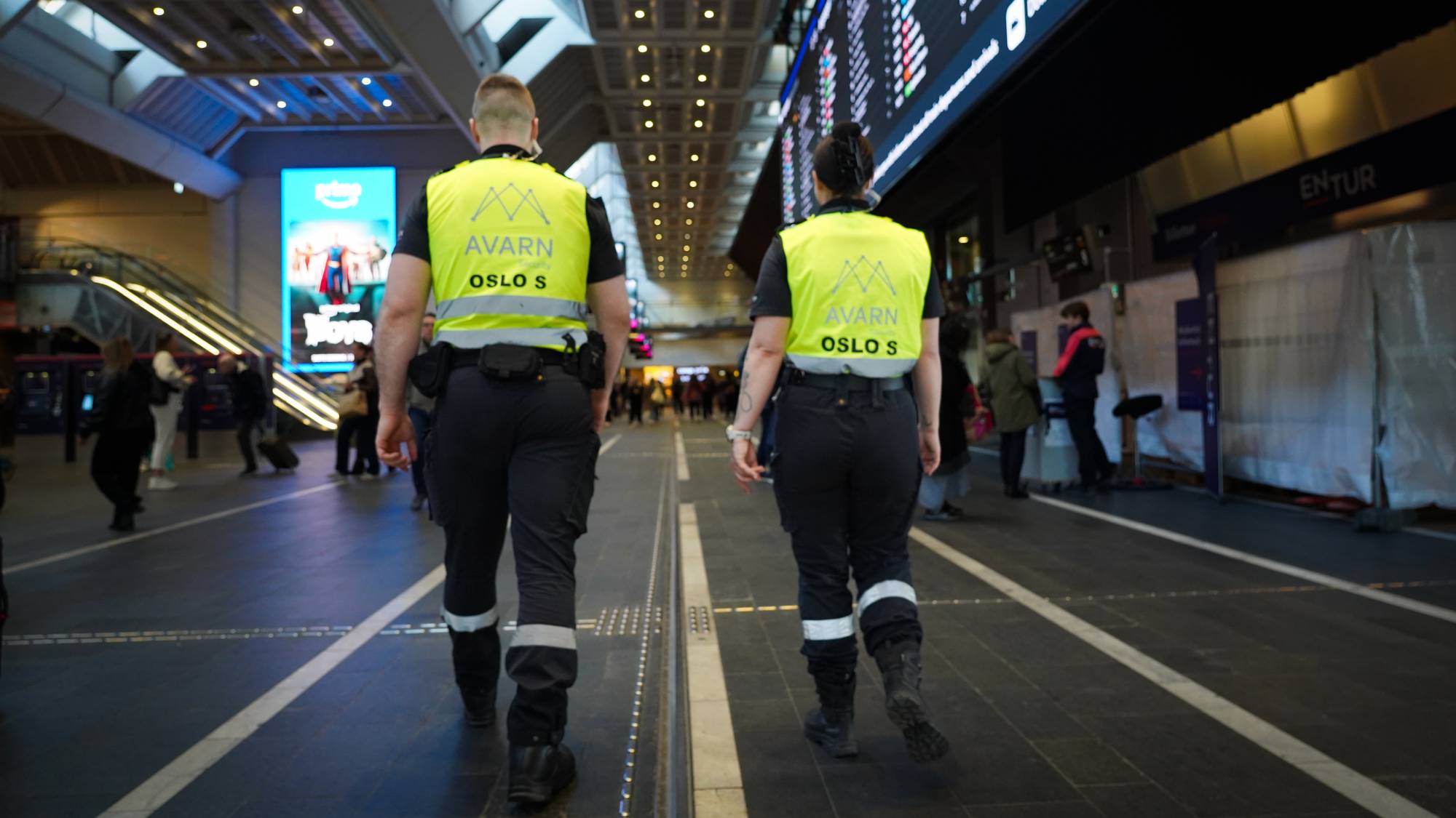 To menn i gule vester med teksten AVARN OSLO S går gjennom en stasjon. De er i forgrunnen av bildet, og bak dem er det flere mennesker. En stor digital tavle med avgangstider henger i bakgrunnen. (Bildebeskrivelsen er laget av en KI-tjeneste)