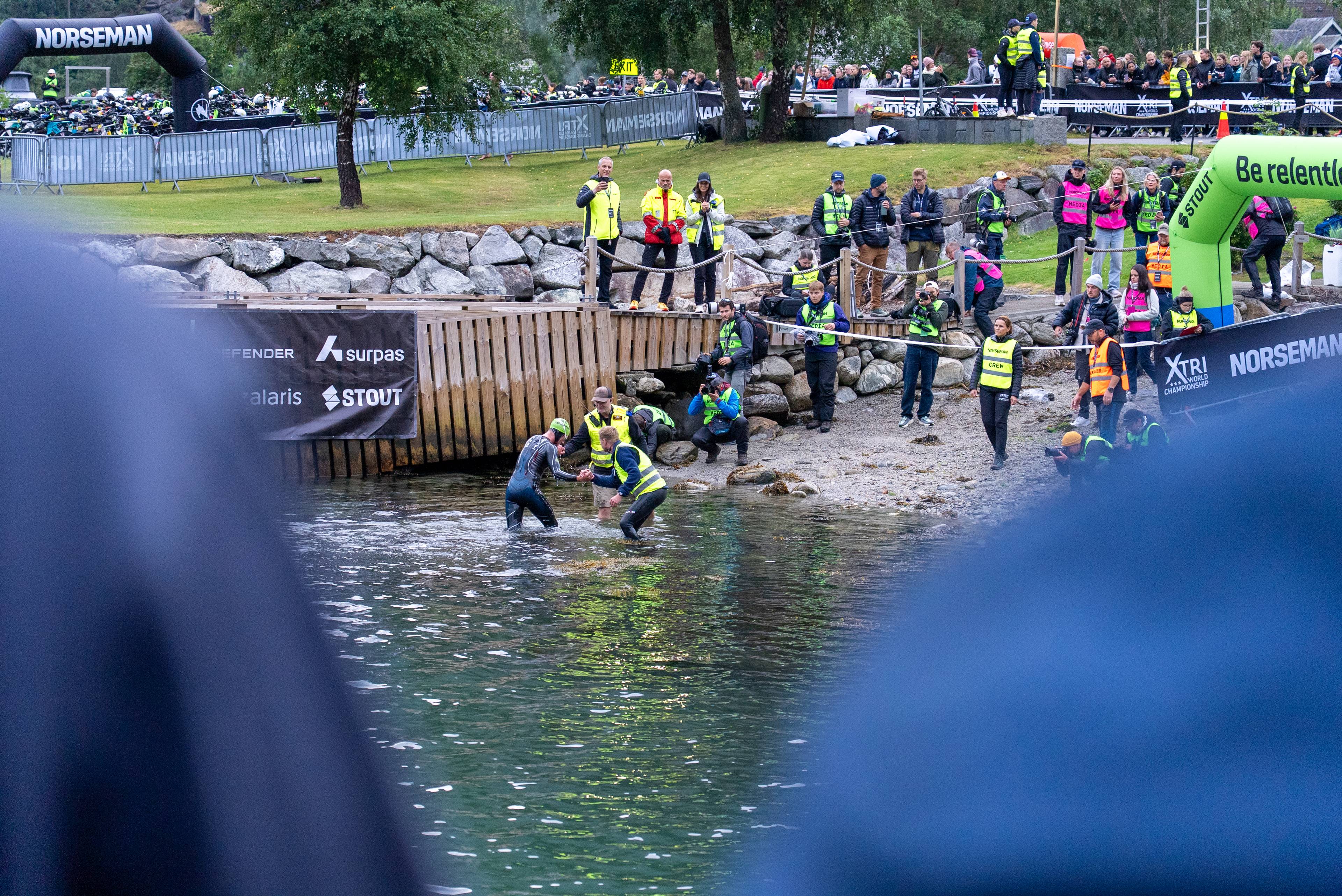 Deltakere går i land fra vannet under Norseman-konkurransen. Funksjonærer og publikum står på land. (Bildebeskrivelsen er laget av en KI-tjeneste)