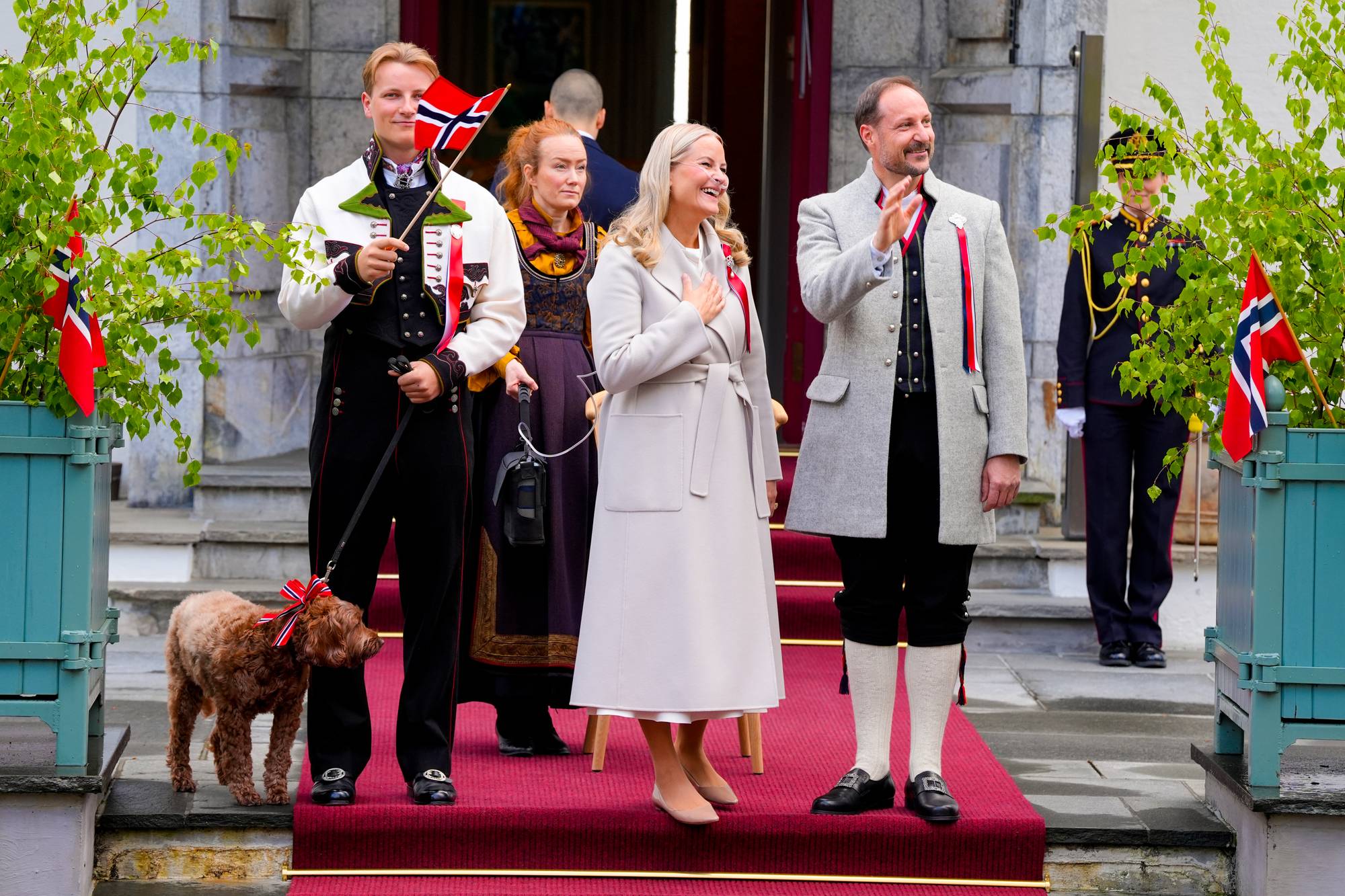 Kronprins Haakon, Kronprinsesse Mette-Marit og Prinsesse Märtha Louise deltar på en markering. Kronprinsesse Mette-Marit har hånden på brystet, mens Kronprins Haakon vinker. En hund med sløyfe står ved siden av Kronprins Haakon. (Bildebeskrivelsen er laget av en KI-tjeneste)