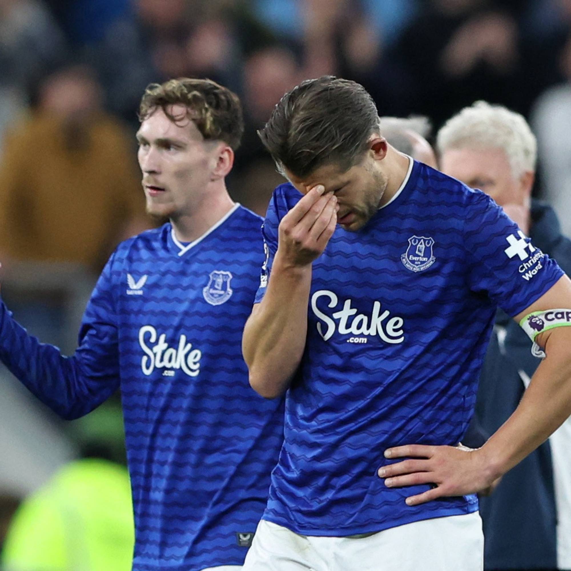 To fotballspillere i blå drakter fra Everton står på en bane. En av spillerne holder seg til ansiktet med hånden, mens den andre ser ut mot siden. Begge spillerne har lagopper. Tilskuerne ses i bakgrunnen. (Bildebeskrivelsen er laget av en KI-tjeneste)