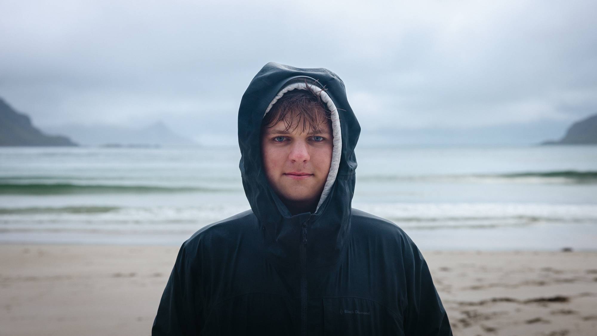 Et portrett av Kristoffer. Han står på en vakker strand i Lofoten med hettegenser og regnjakke, begge hettene på. Han ser rett i kamera, våt i håret. Det ser friskt og kaldt ut.