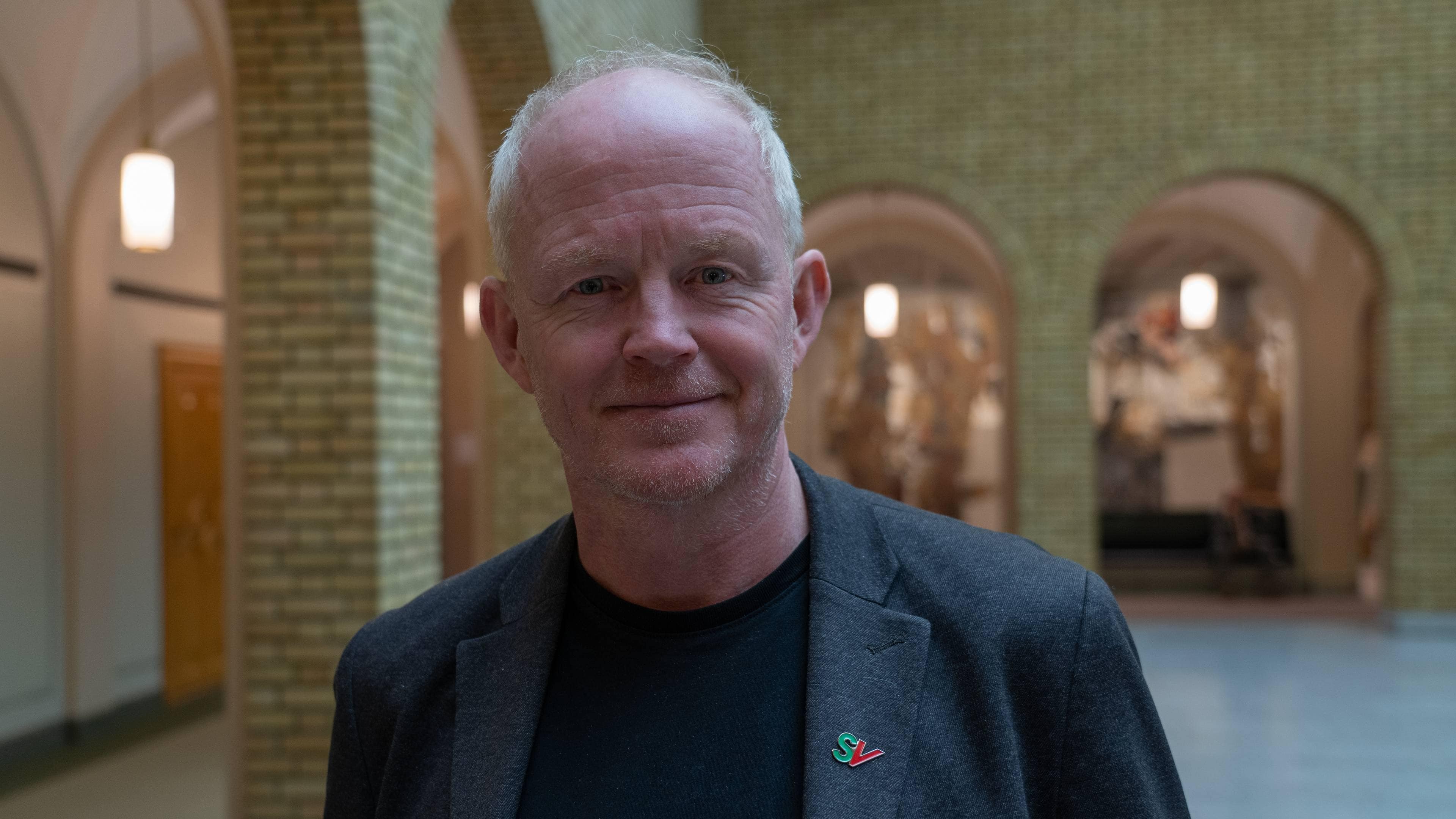 Lars Haltbrekken står i en romslig bygning med mursteinsvegger, iført en grå blazer og svart t-skjorte. Han har et pin-merke med teksten "SV" festet på jakken. Omgivelsene har moderne kunstverk synlige i bakgrunnen, som kan indikere et offentlig eller kulturelt arrangement. Belysningen i rommet er diffus og skaper en åpen atmosfære. (Bildebeskrivelsen er laget av en KI-tjeneste)