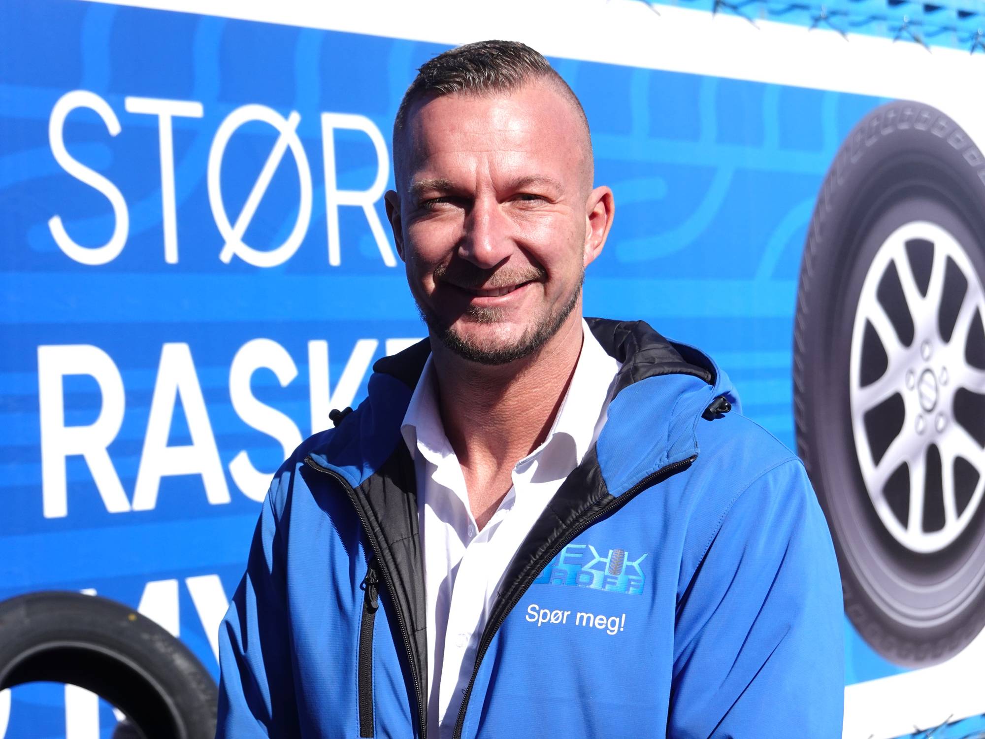 En mann i blå jakke smiler foran en blå bakgrunn med hvit tekst. På jakken er et logo med teksten "DEKK PROFF". Bakgrunnen viser et dekk og hjul. Mannen har kortklipt hår. (Bildebeskrivelsen er laget av en KI-tjeneste)