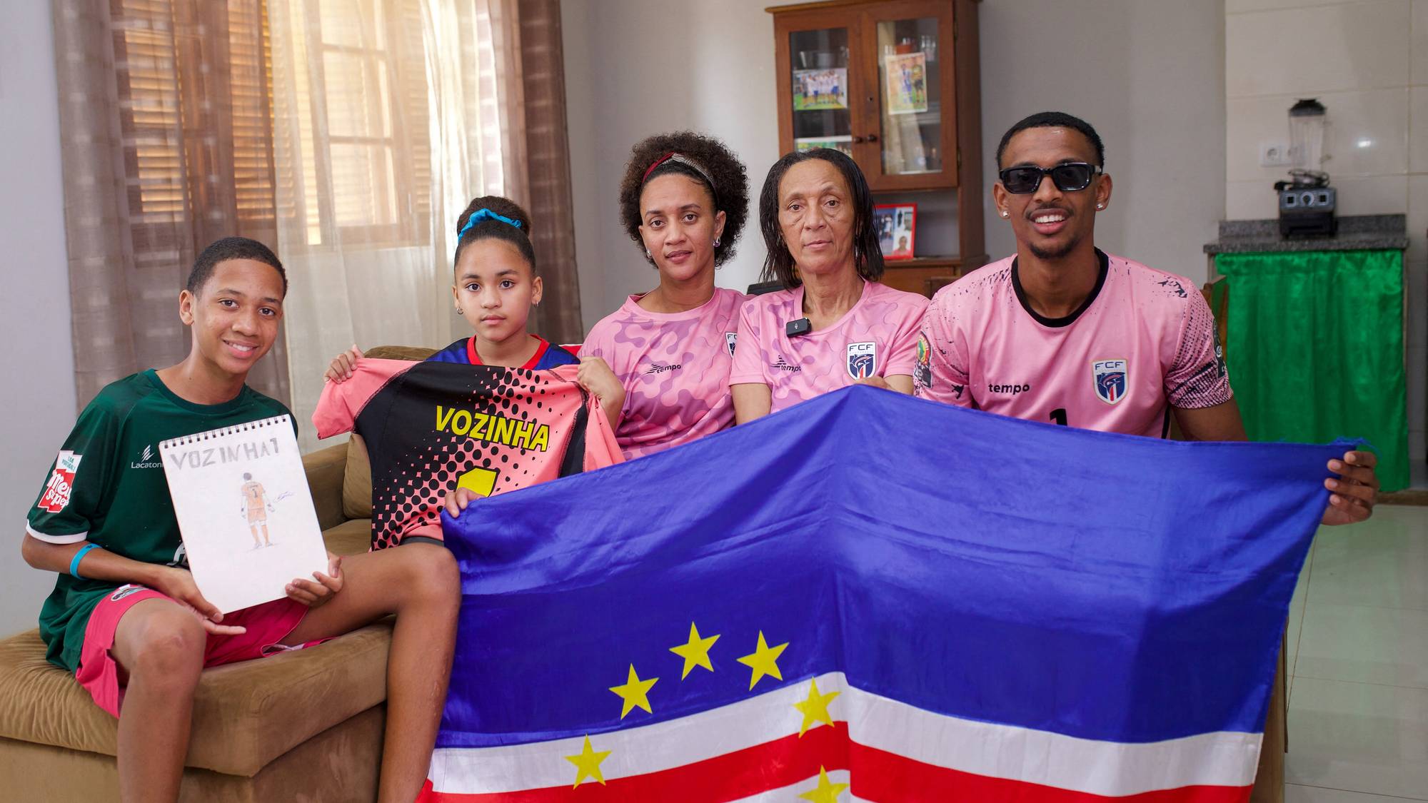 En familie holder et flagg fra Kapp Verde. En gutt holder et tegnet portrett av en person. Flere av familiemedlemmene har på seg rosa fotballdrakter. (Bildebeskrivelsen er laget av en KI-tjeneste)