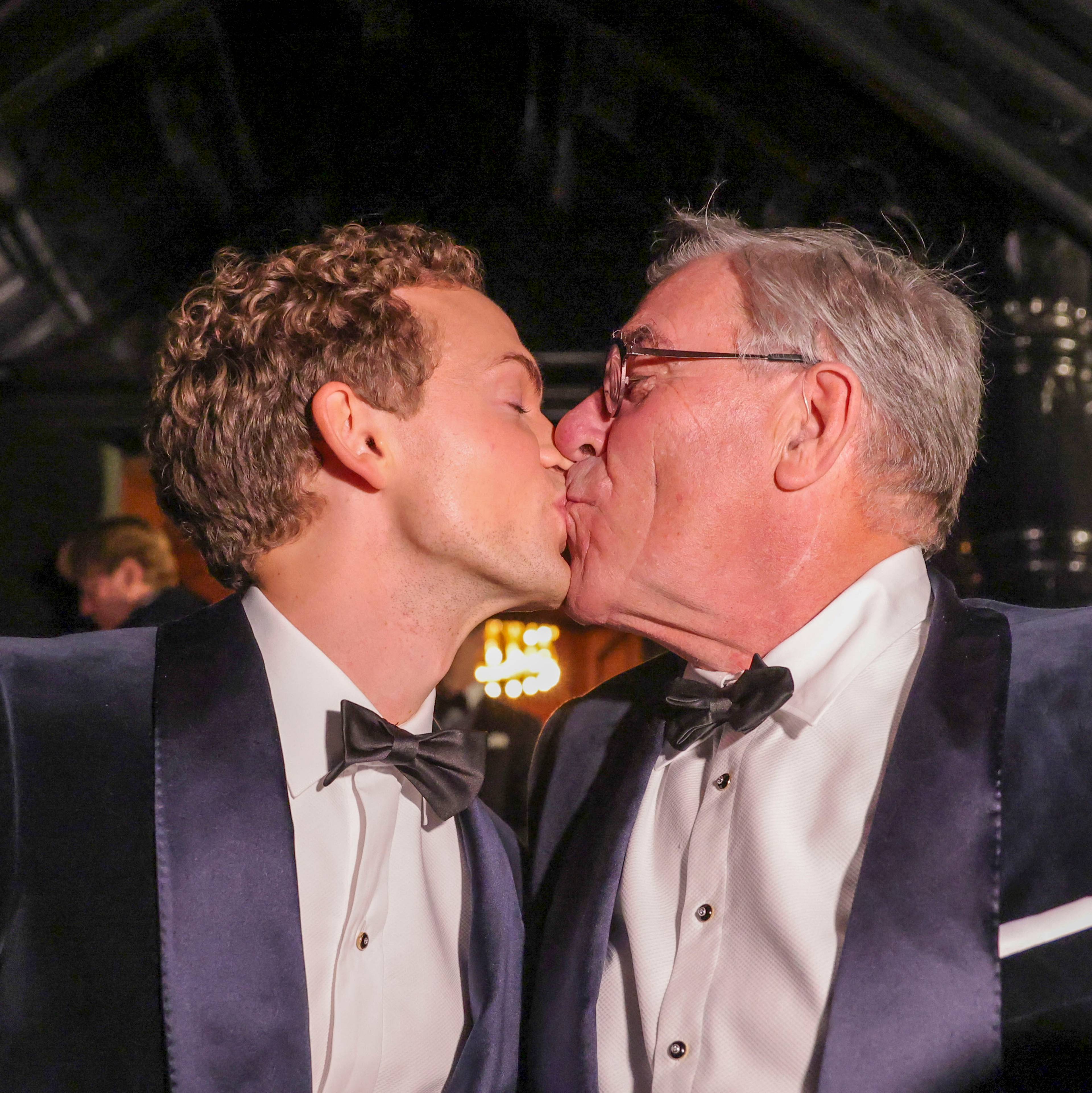 To men in formal wear kiss. They are holding hands. The man on the left has curly hair and is wearing a tuxedo. The man on the right is older and wearing glasses. The background is dark and out of focus. (Bildebeskrivelsen er laget av en KI-tjeneste)