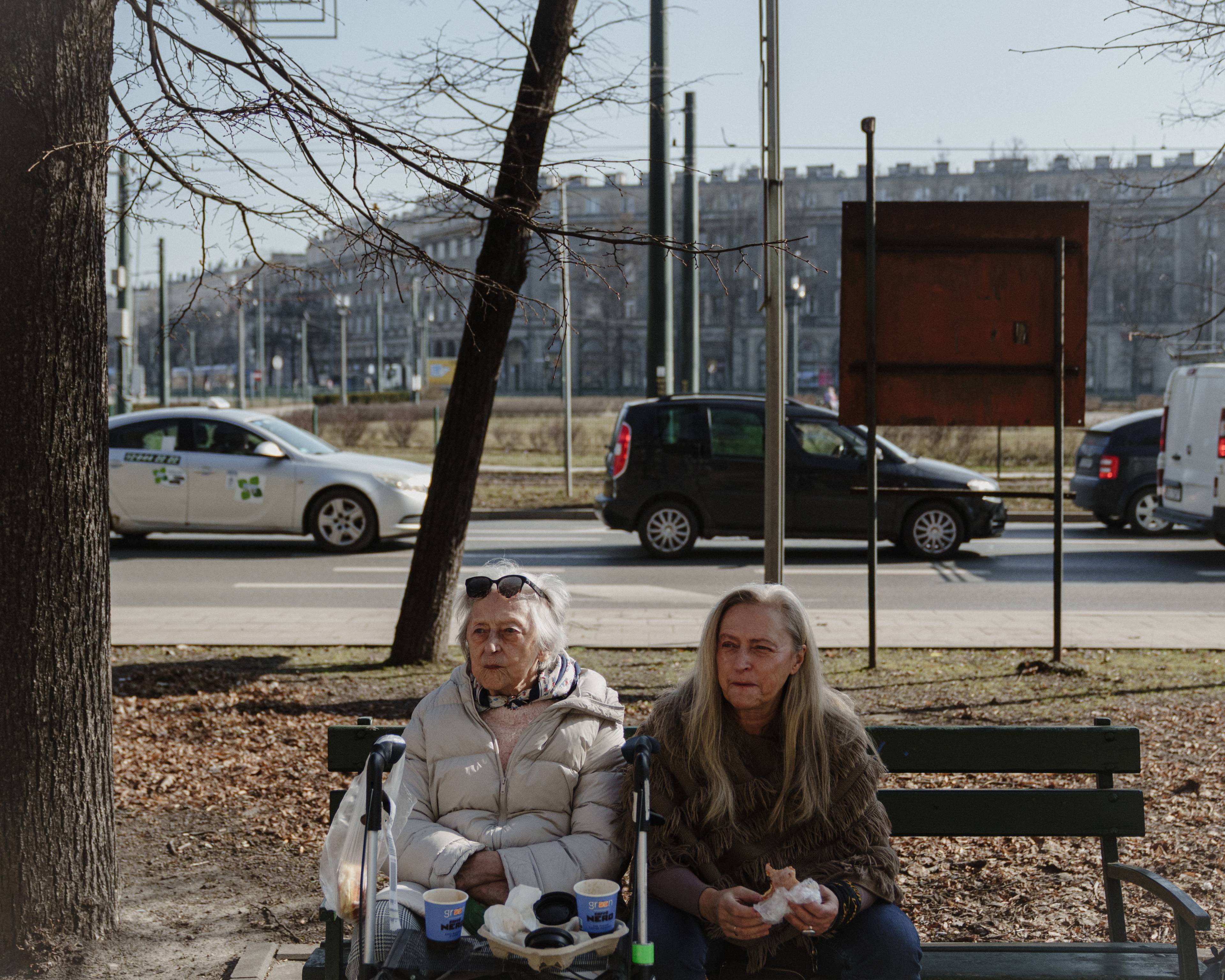 Magdalena, en gammel kvinne med hvitt hår, sitter ved siden av Edith, en gammel, men noe yngre kvinne med grått hår. De sitter på en benk foran en bilvei i en by