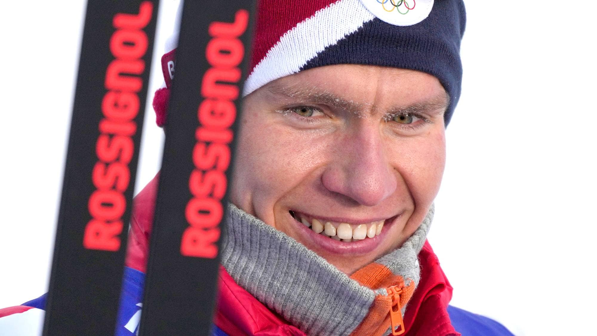 Nærstudiebilde av en mannlig alpinist med ski. Han smiler og har på seg en lue med olympiske ringer. Luen er blå, rød og hvit. Mannen har på seg en blå og rød jakke med grå krage. Skismittet er sorte med rød tekst. (Bildebeskrivelsen er laget av en KI-tjeneste)