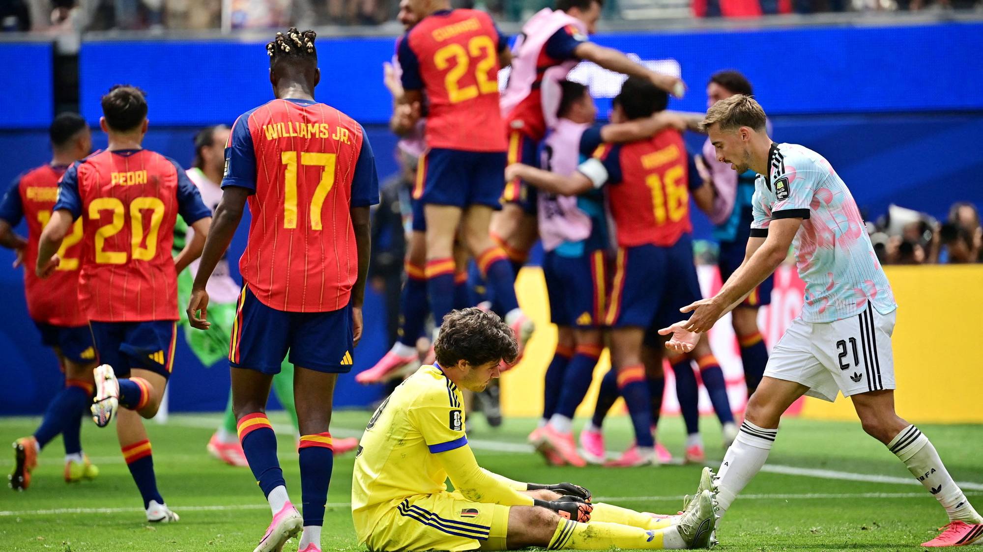 Fotballspillere i røde drakter feirer mens en sittende keeper i gult og en motspiller ser på. (Bildebeskrivelsen er laget av en KI-tjeneste)