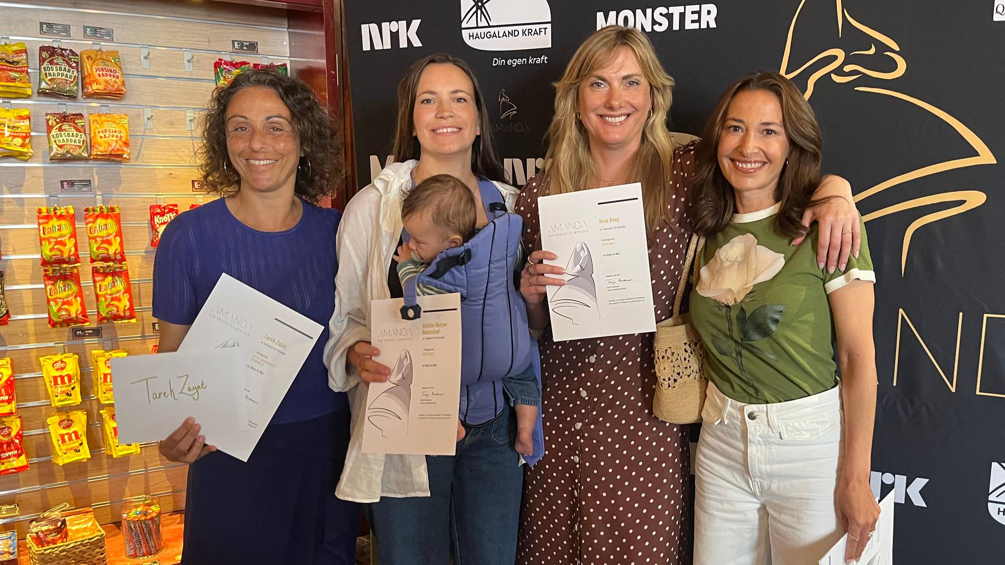 Fire kvinner står og smiler til kameraet foran en svart bakvegg med sponsorlogoer. På venstre flanke står Pia Tjelta. To av kvinnene holder utdelte diplomer. En av kvinnene bærer et spedbarn i bæresjal. Bak kvinnene er det hyller med godteri. (Bildebeskrivelsen er laget av en KI-tjeneste)